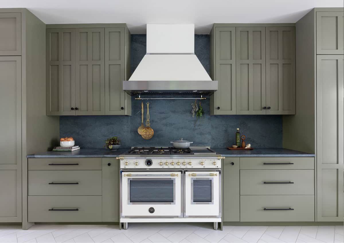 A white Bertazzoni dual-oven range with brass trim under a white hood in an olive green kitchen with a blue stone backsplash