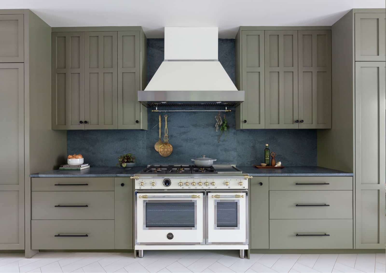 A white Bertazzoni dual-oven range with brass trim under a white hood in an olive green kitchen with a blue stone backsplash