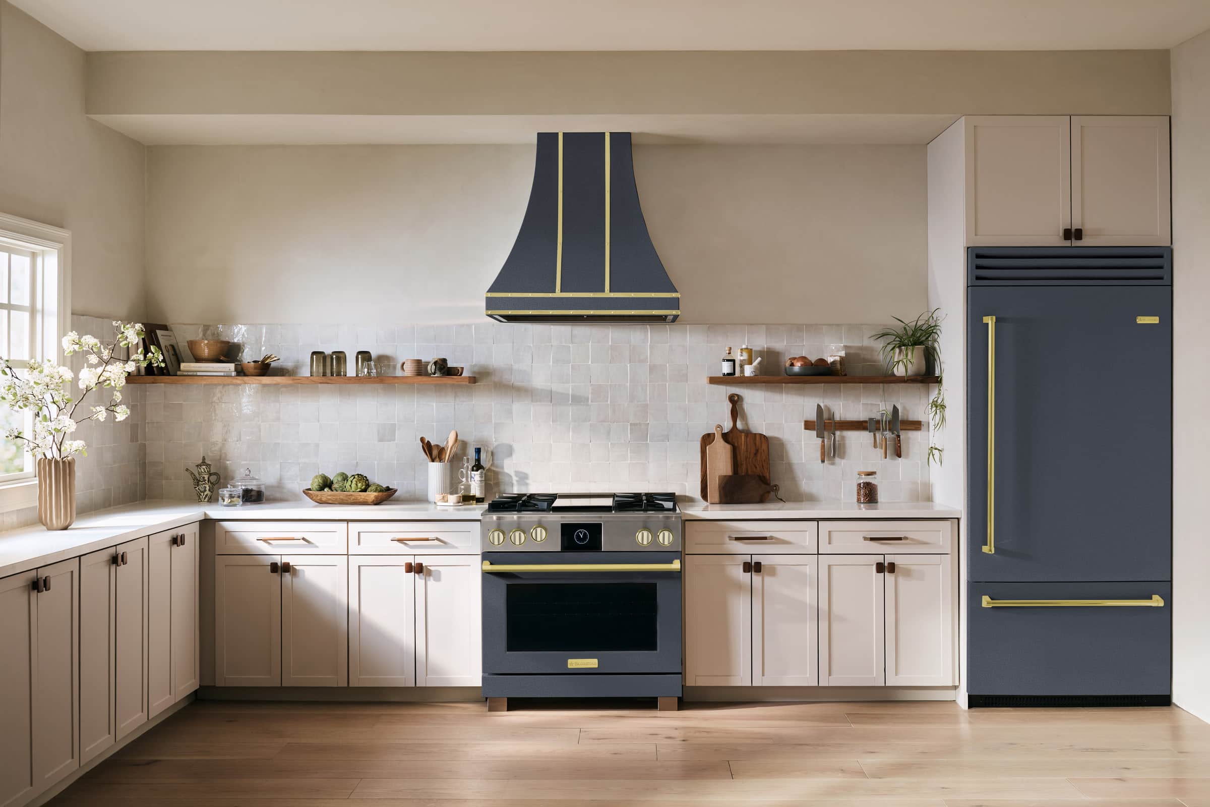 A bluestone BlueStar range with brushed brass trim under a matching hood, beside a bluestone built-in refrigerator with a bottom freezer drawer, in a cream kitchen with open wood shelves