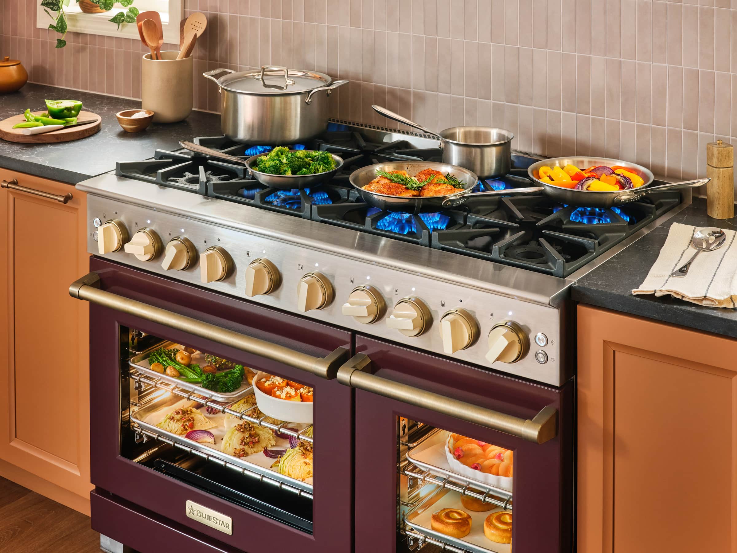 A burgundy 48-inch BlueStar range with cream knobs on brass bezels, its open burners lit under pans of vegetables and chicken, and trays of vegetables and pastries lit behind both closed oven doors
