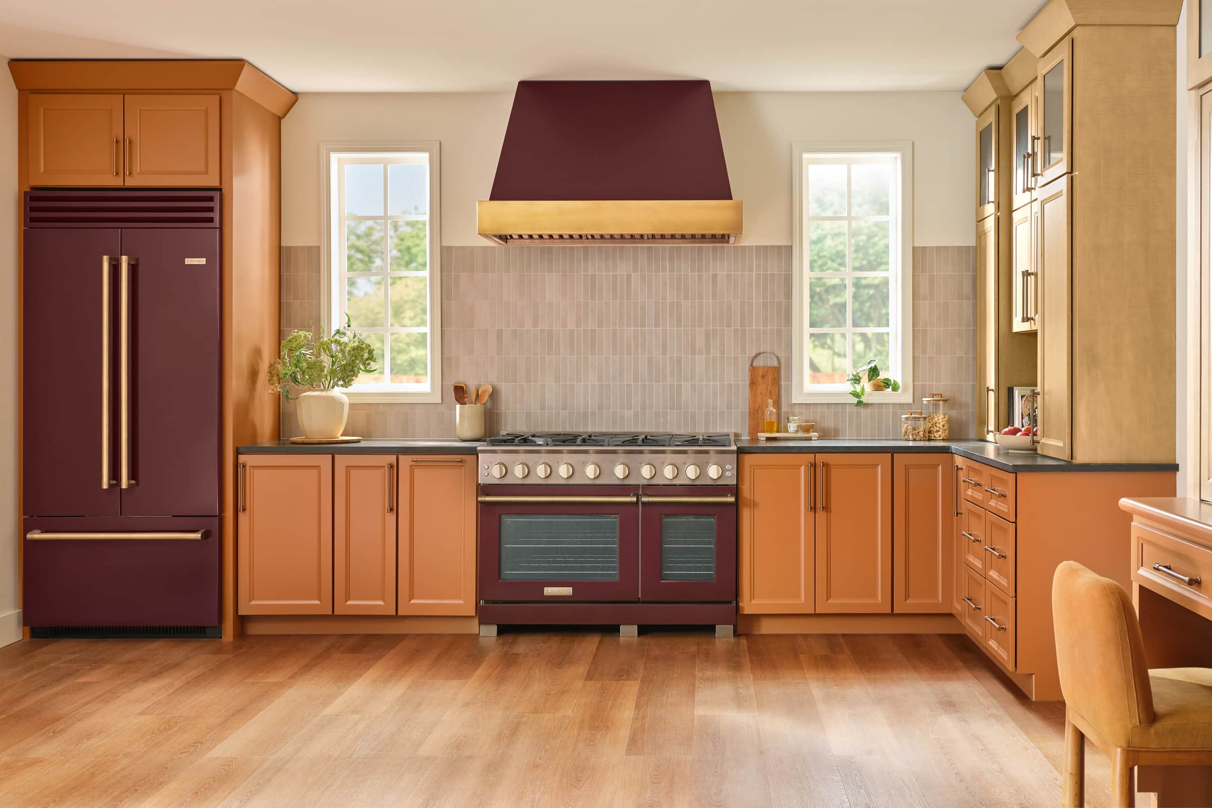 A burgundy 48-inch BlueStar range under a burgundy and brass hood, with a matching French-door refrigerator, in a kitchen with terracotta cabinets