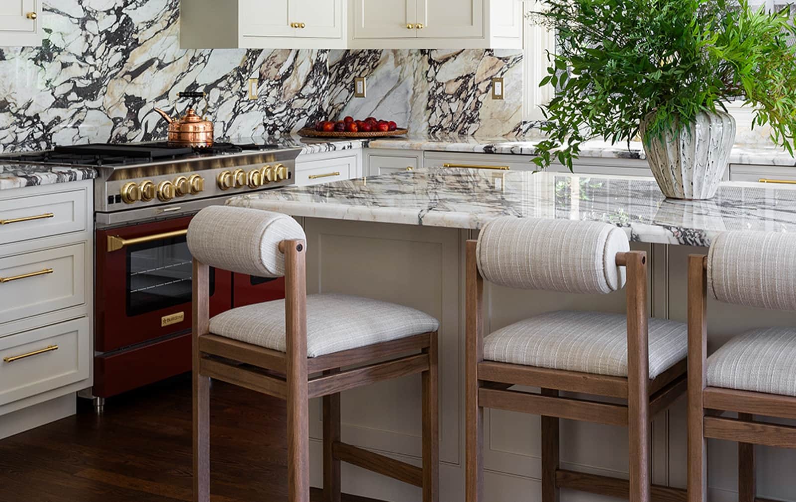 A deep red BlueStar range with brass trim and a marble backsplash beside a marble-topped island in a cream kitchen