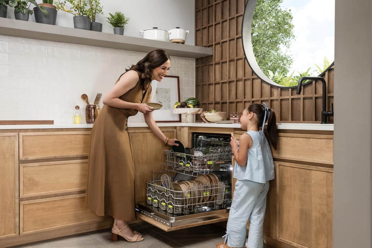 An open Cove dishwasher being loaded in a light wood kitchen with a round window and shelves of potted herbs