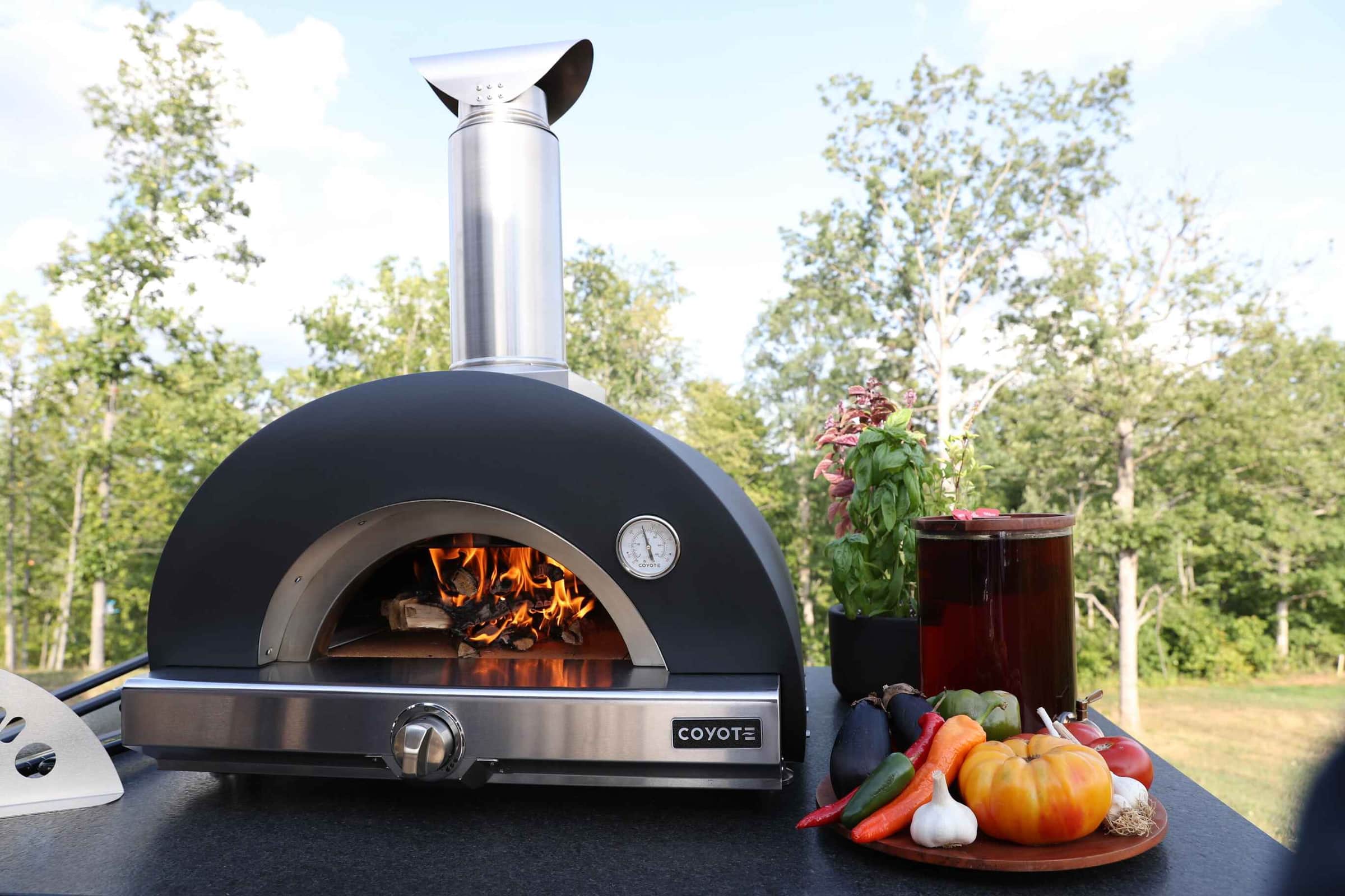 A black Coyote wood-fired pizza oven with a live fire beside a bowl of vegetables outdoors