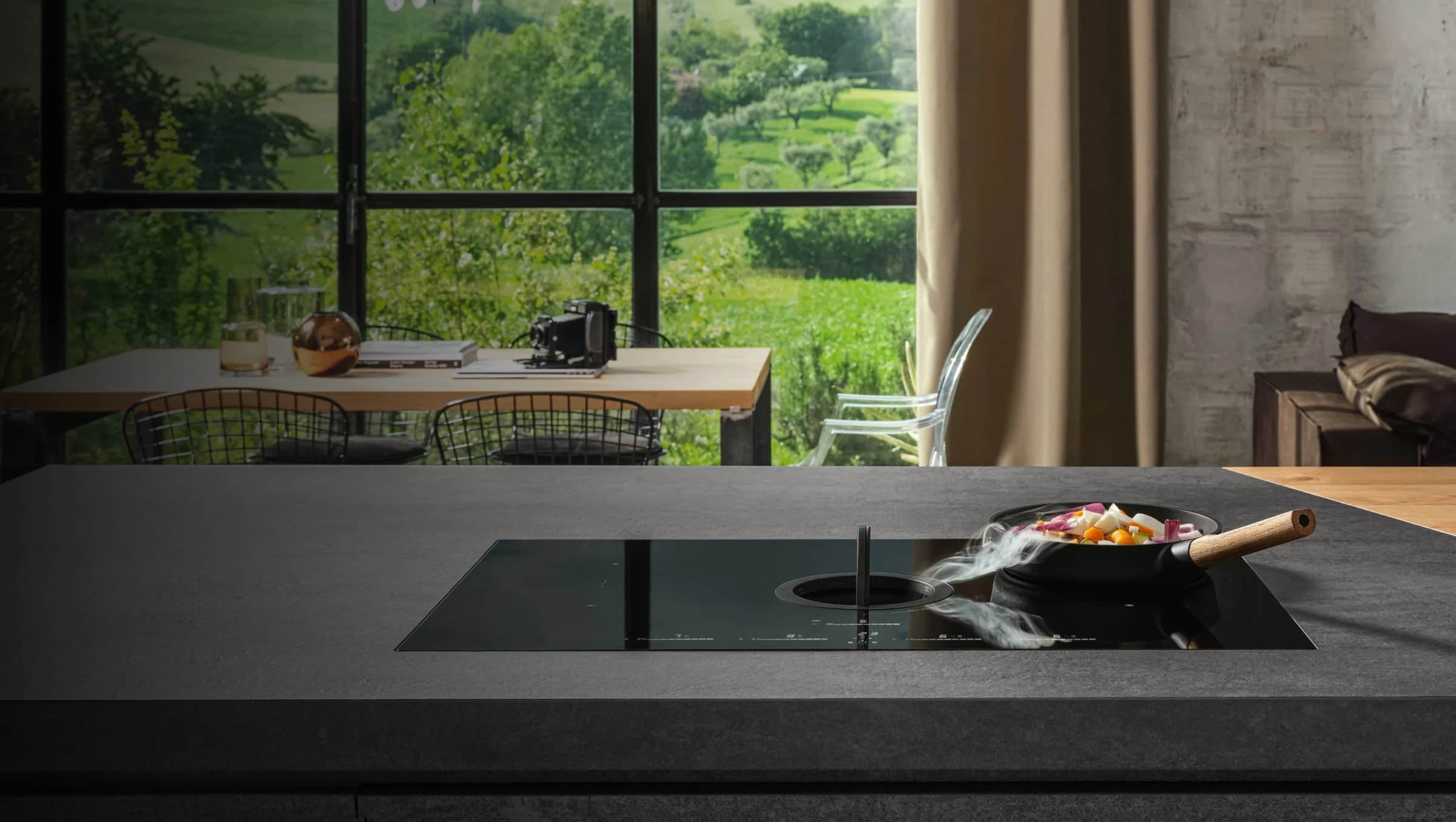 A black Elica induction cooktop with a central round downdraft vent set into a dark stone island, steam from a pan of vegetables drawn into the vent, tall windows onto green hills behind