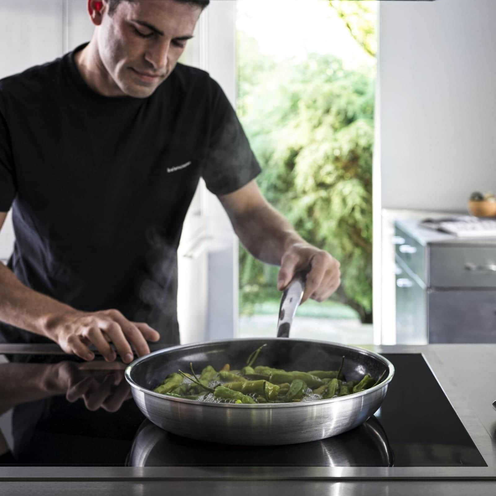 A chef searing shishito peppers in a stainless pan on a Gaggenau induction cooktop