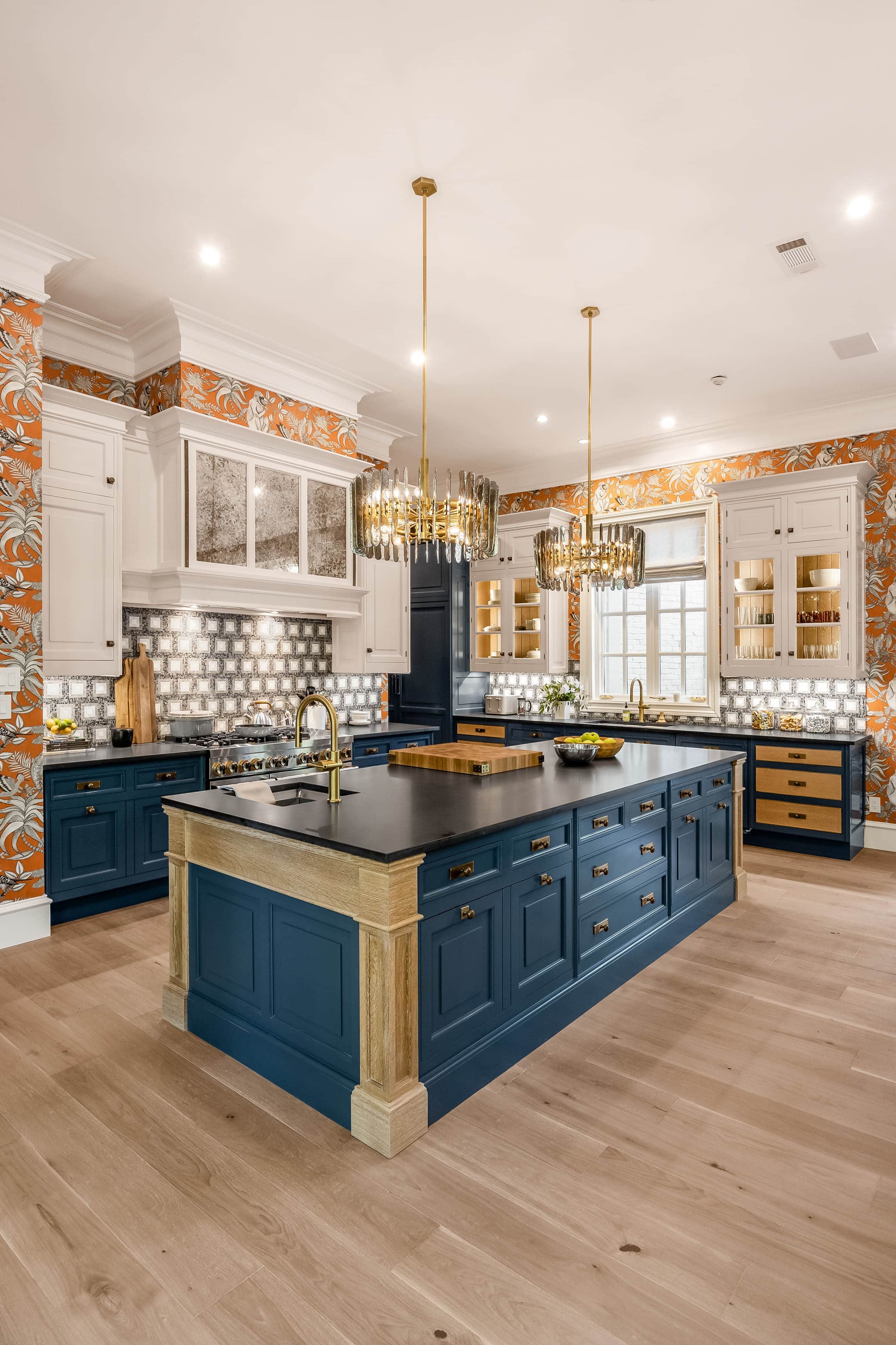 A blue kitchen island under brass chandeliers with JennAir appliances against floral wallpaper