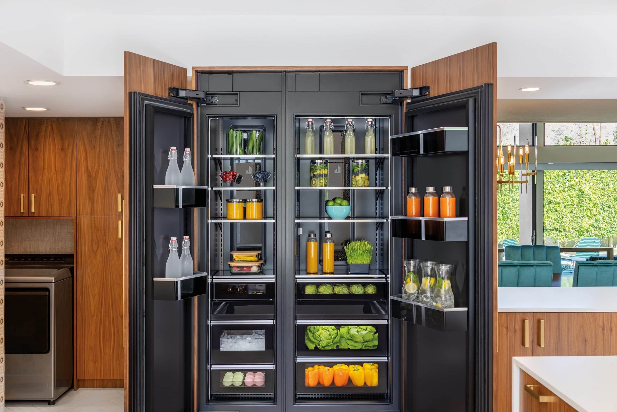 Two JennAir column refrigerators with walnut panel doors standing open on lit black interiors stocked with produce and bottled drinks