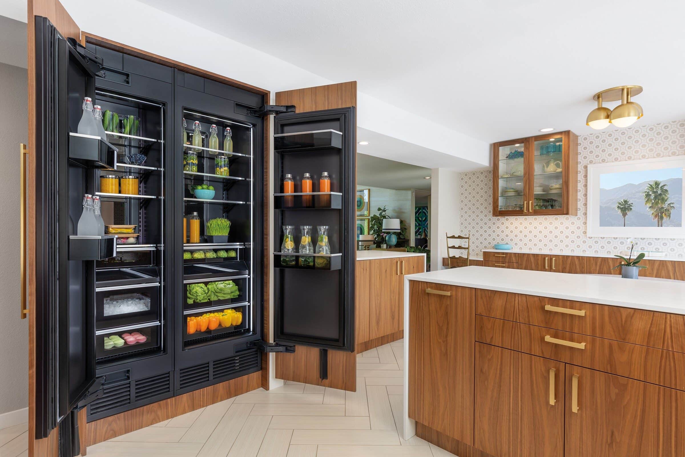 Two built-in JennAir column refrigerators behind walnut cabinet panels, both doors open on lit black interiors stocked with produce and glass bottles, beside a walnut island
