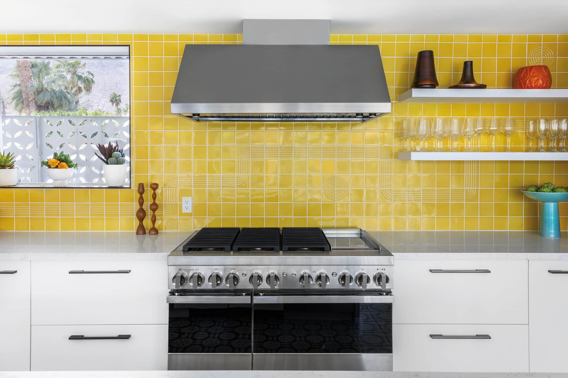 A stainless JennAir professional range with a griddle under a stainless chimney hood, set against a wall of glossy yellow tile with white floating shelves of glassware