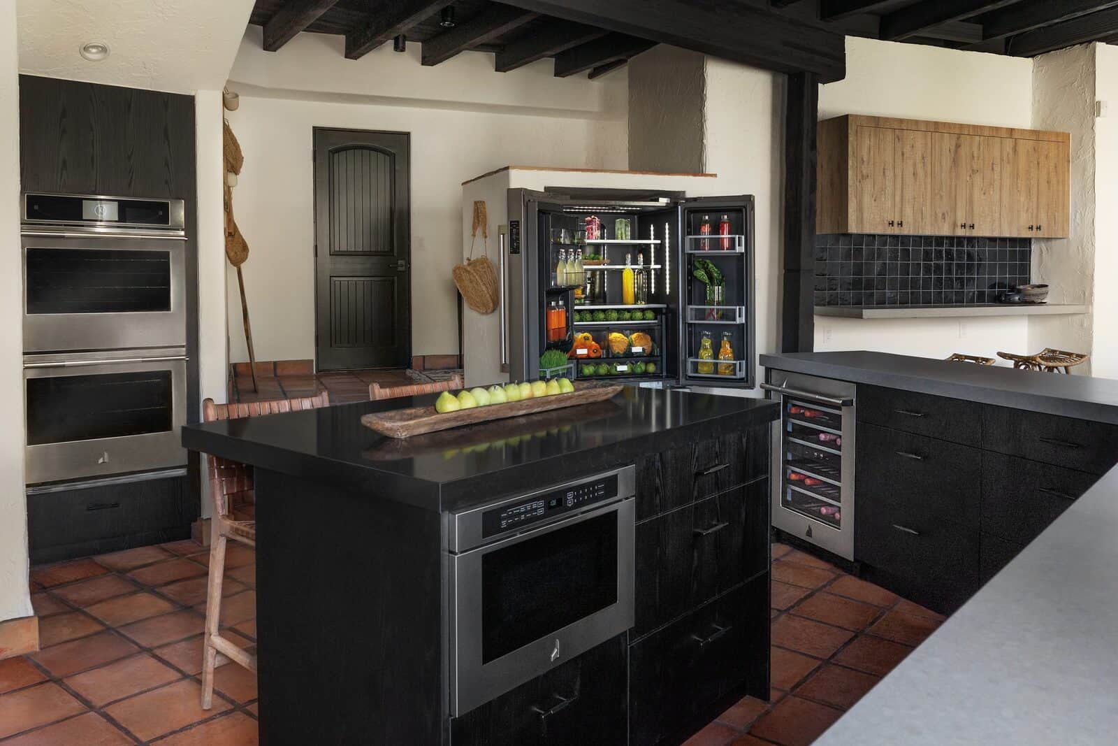 A Spanish-style kitchen with a black island housing a JennAir microwave drawer, stainless double wall ovens, an open black-stainless JennAir refrigerator, and a terracotta tile floor under dark ceiling beams