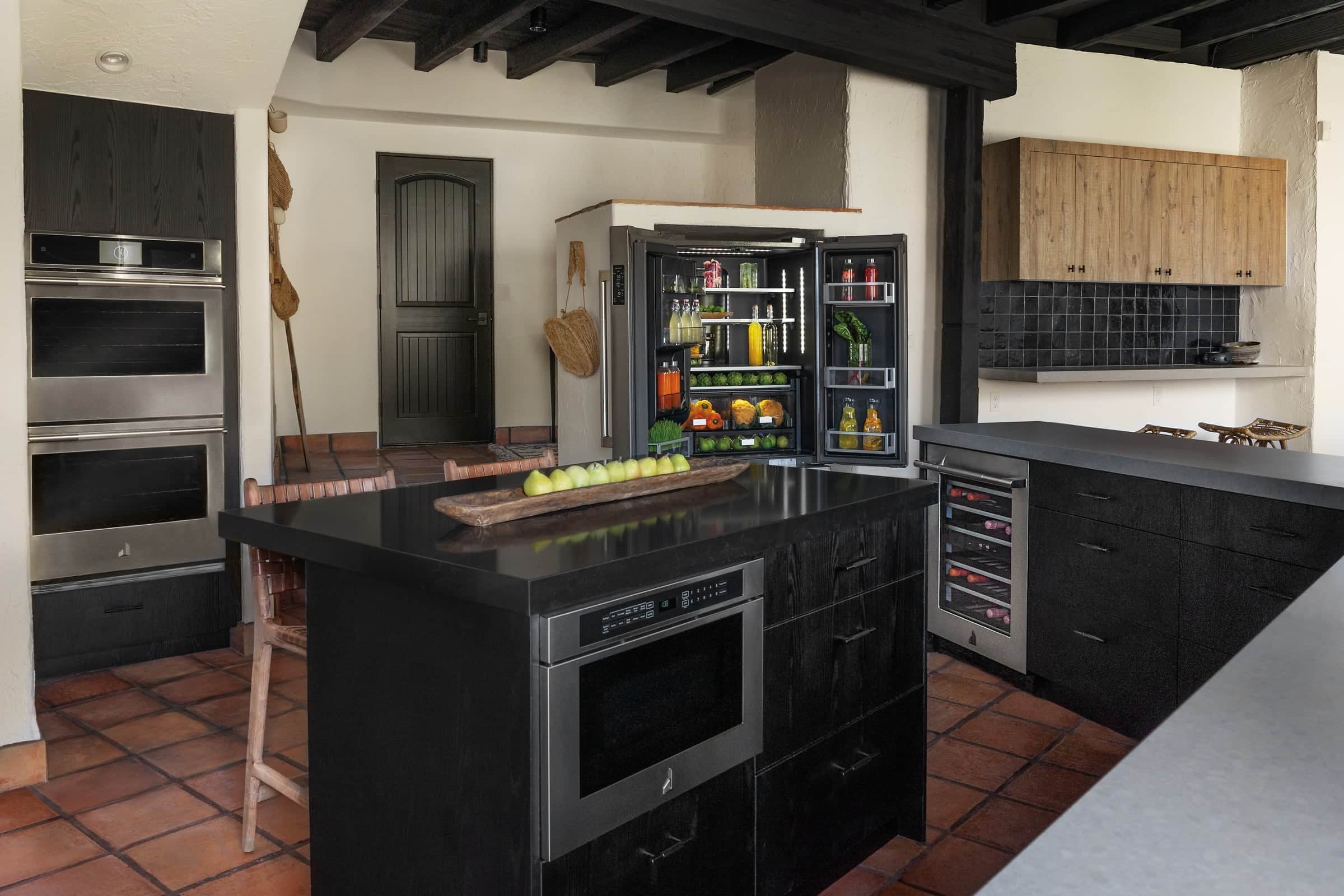 A Spanish-style kitchen with a black island housing a JennAir microwave drawer, stainless double wall ovens, an open black-stainless JennAir refrigerator, and a terracotta tile floor under dark ceiling beams
