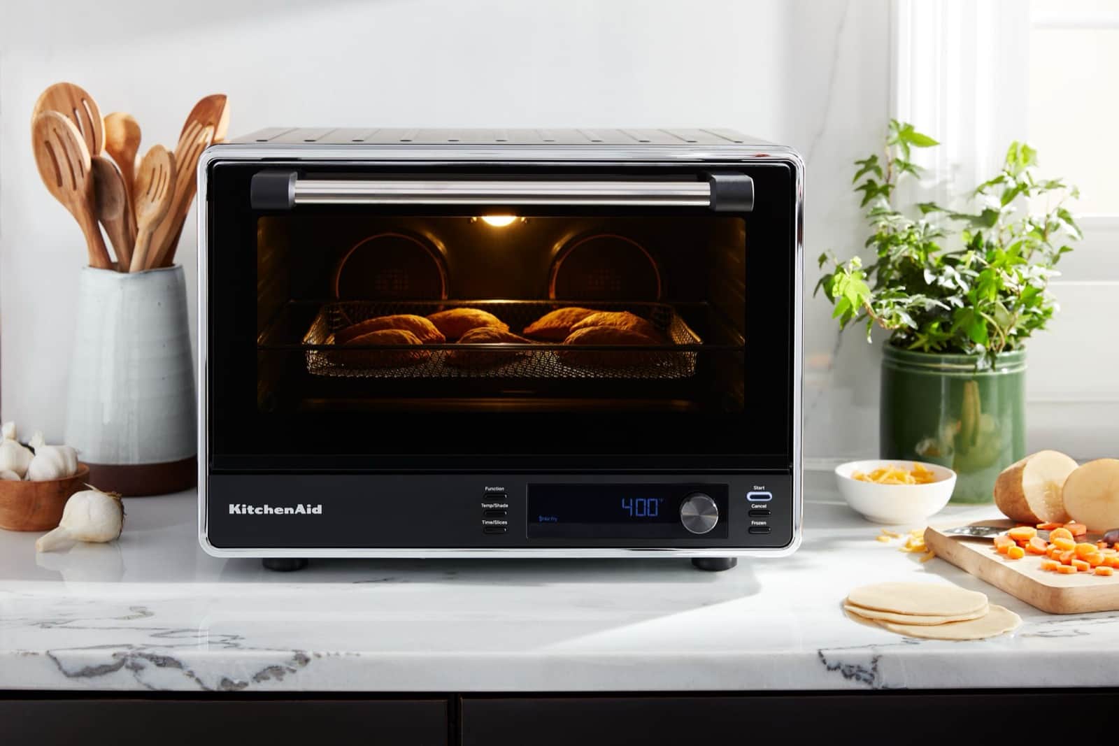 A black KitchenAid countertop oven air-frying hand pies on a marble counter beside fresh vegetables and wood utensils