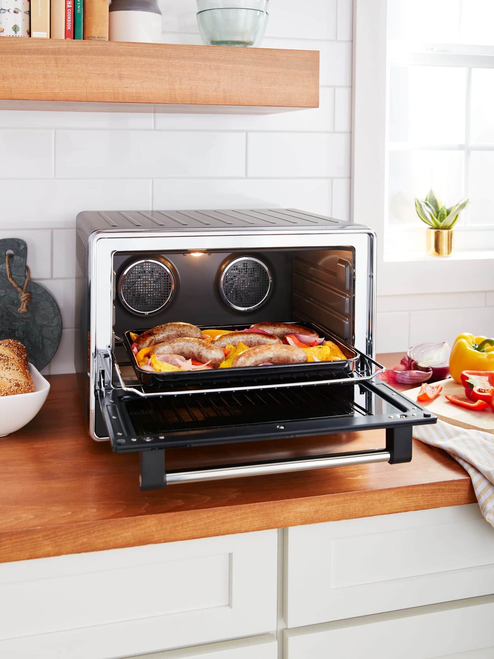 A black KitchenAid countertop oven with its door down on a butcher-block counter, a tray of sausages and sliced peppers on the rack, white subway tile behind