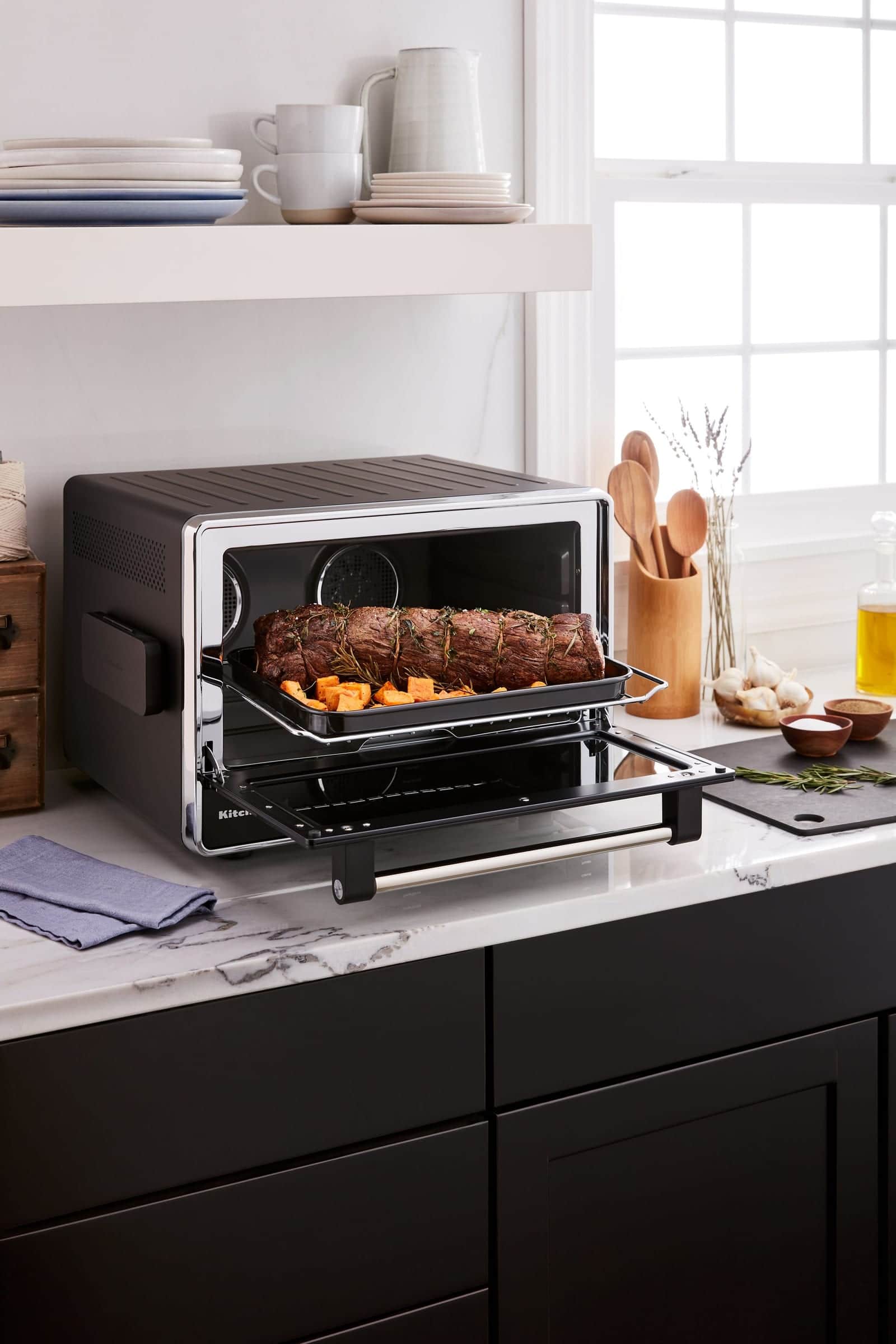 A black KitchenAid countertop oven with its door open on a tied roast and sweet potatoes, set on a white marble counter above black cabinets