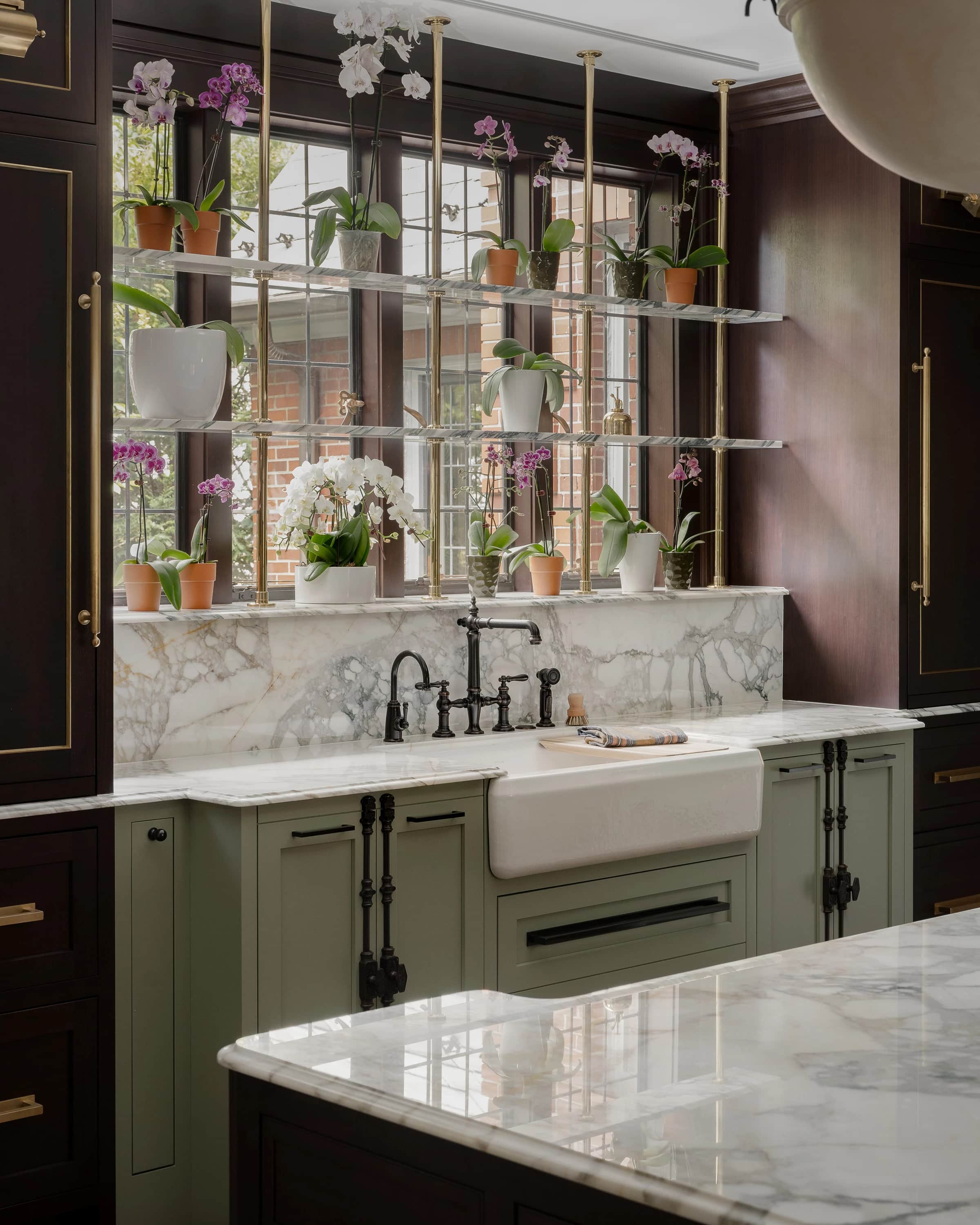 A white Kohler apron-front sink with a bridge faucet in olive cabinetry under shelves of orchids