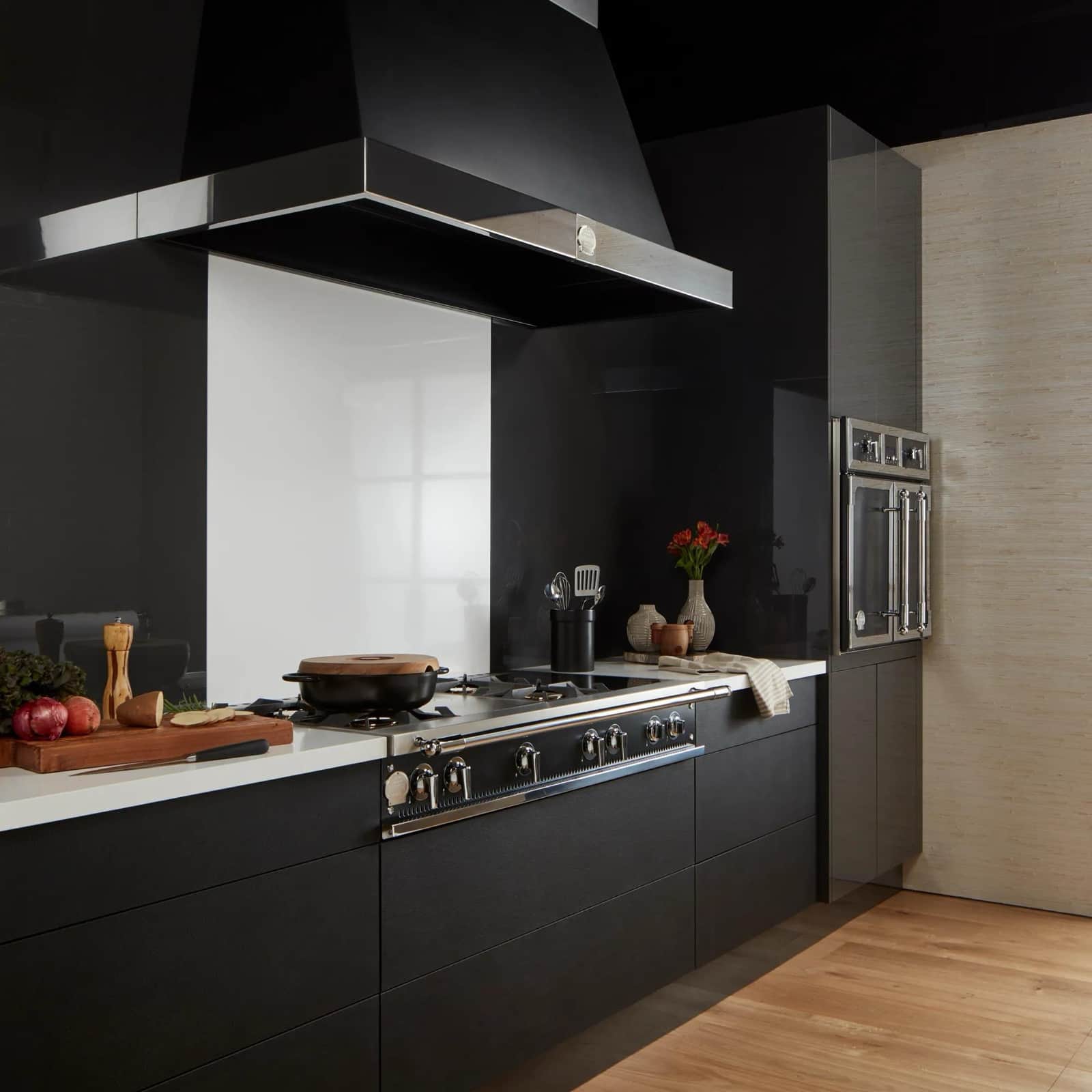 A stainless La Cornue cooktop and matching wall hood in a matte black kitchen with an oak floor and a built-in wall oven