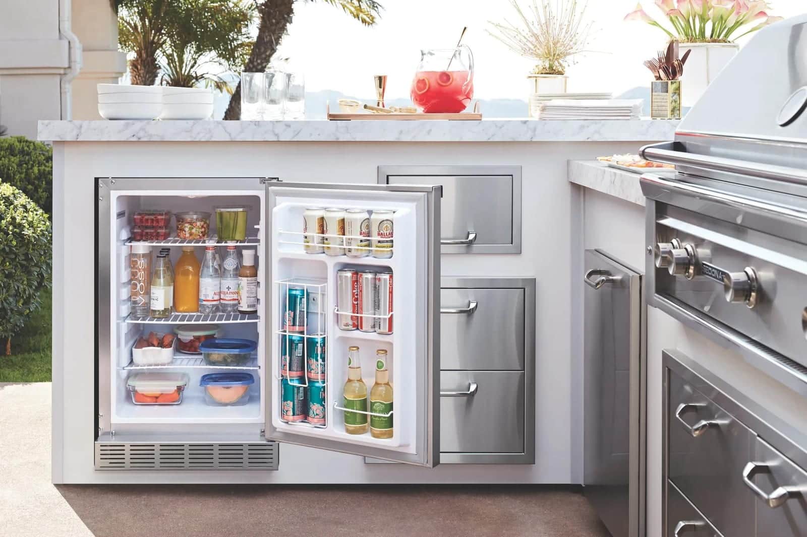 An open stainless Lynx outdoor undercounter refrigerator holding drinks and produce, built into a white marble-topped island beside garden greenery