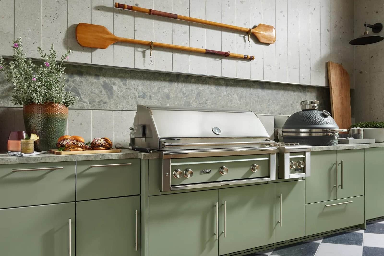 A sage green outdoor kitchen with a built-in stainless Lynx grill, a side burner, and a ceramic kamado grill under a stone-tiled wall hung with wooden pizza peels
