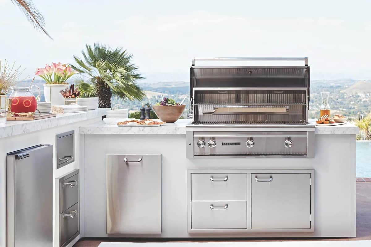 A white outdoor kitchen island with an open stainless Lynx grill, storage drawers, and an undercounter refrigerator on a hillside terrace with palms and a distant valley view