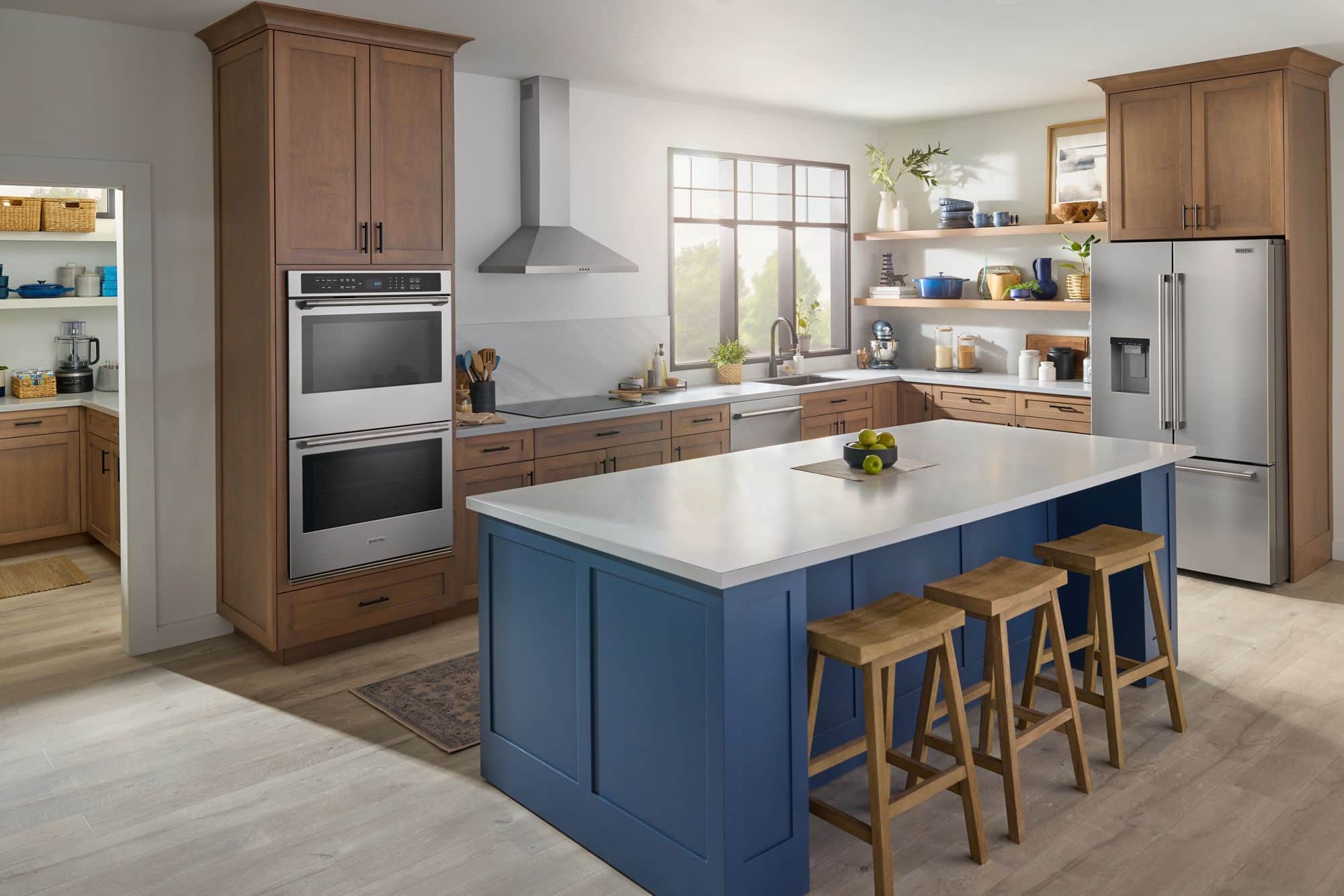 A wide kitchen with a stainless Maytag double wall oven set into wood cabinetry, a blue island with three wood stools, and a stainless French-door refrigerator at the right