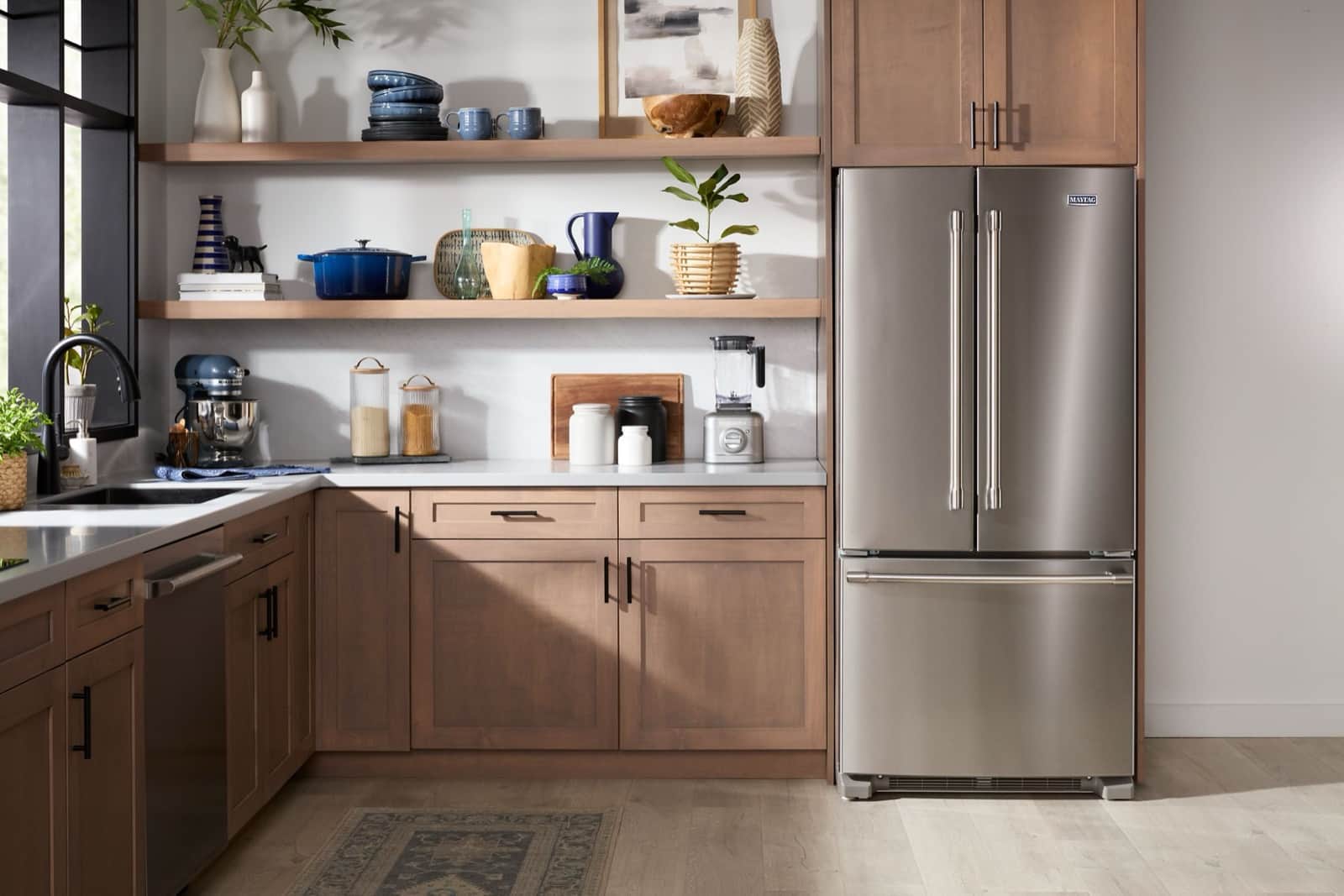 A stainless Maytag French-door refrigerator with a bottom freezer in a warm wood kitchen with open shelves