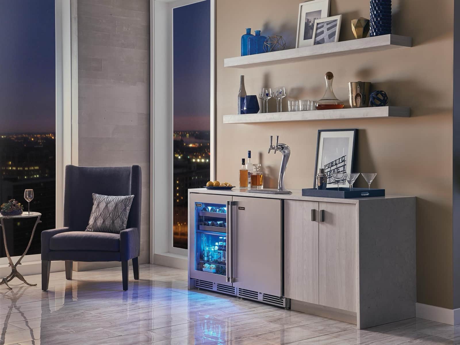 A Perlick stainless glass-door beverage center and tap system built into a home bar cabinet by a window with a city view at dusk