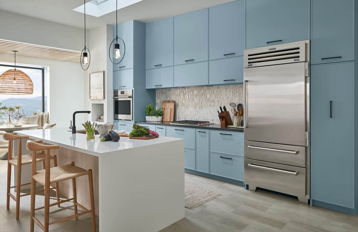 Blue kitchen with a tall stainless Sub-Zero column refrigerator; a Wolf wall oven and gas cooktop; and a white quartz island with fresh vegetables on a cutting board