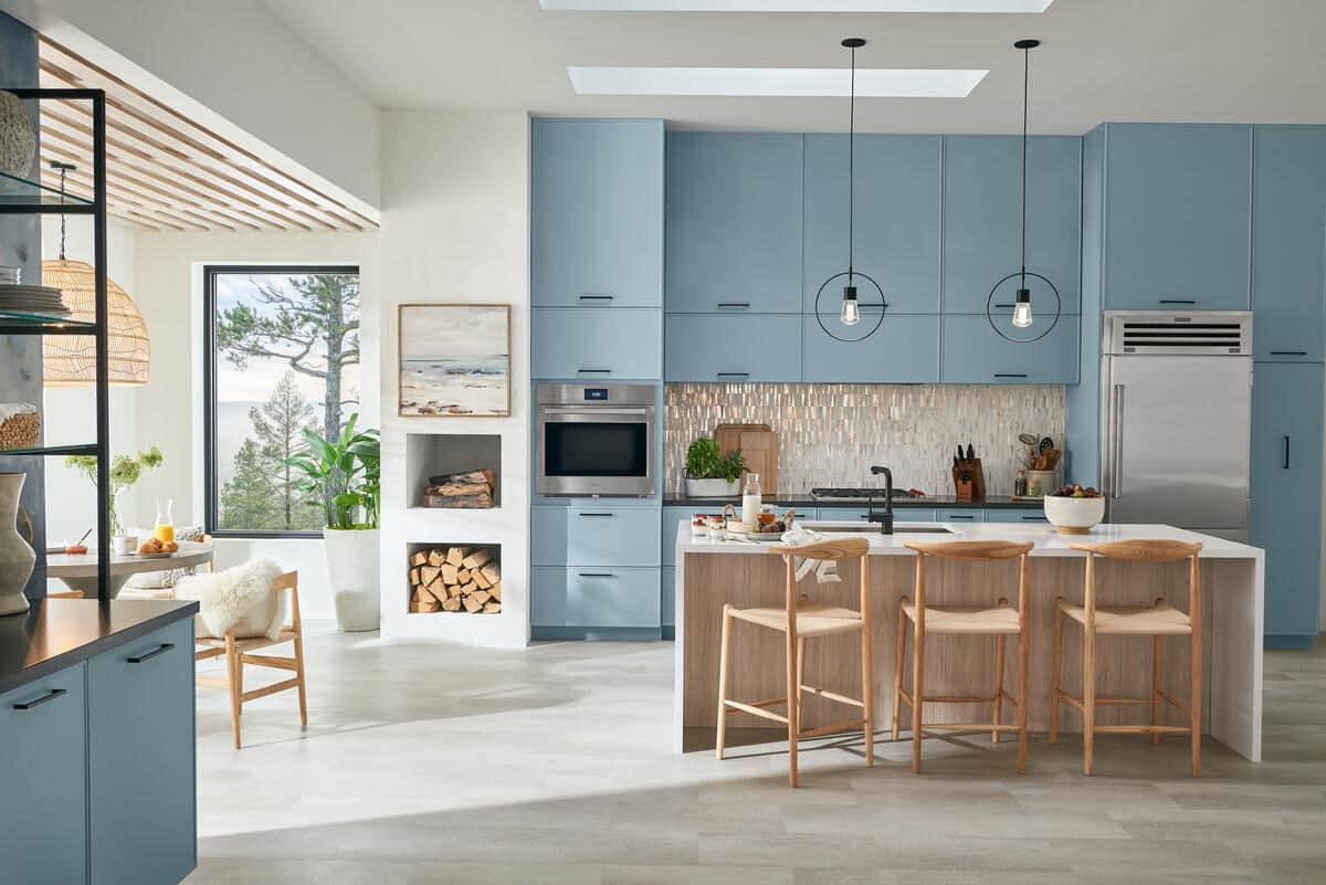 Open-plan kitchen with pale blue cabinetry; a stainless Sub-Zero column refrigerator; a Wolf wall oven and microwave; and a wood island with three woven stools