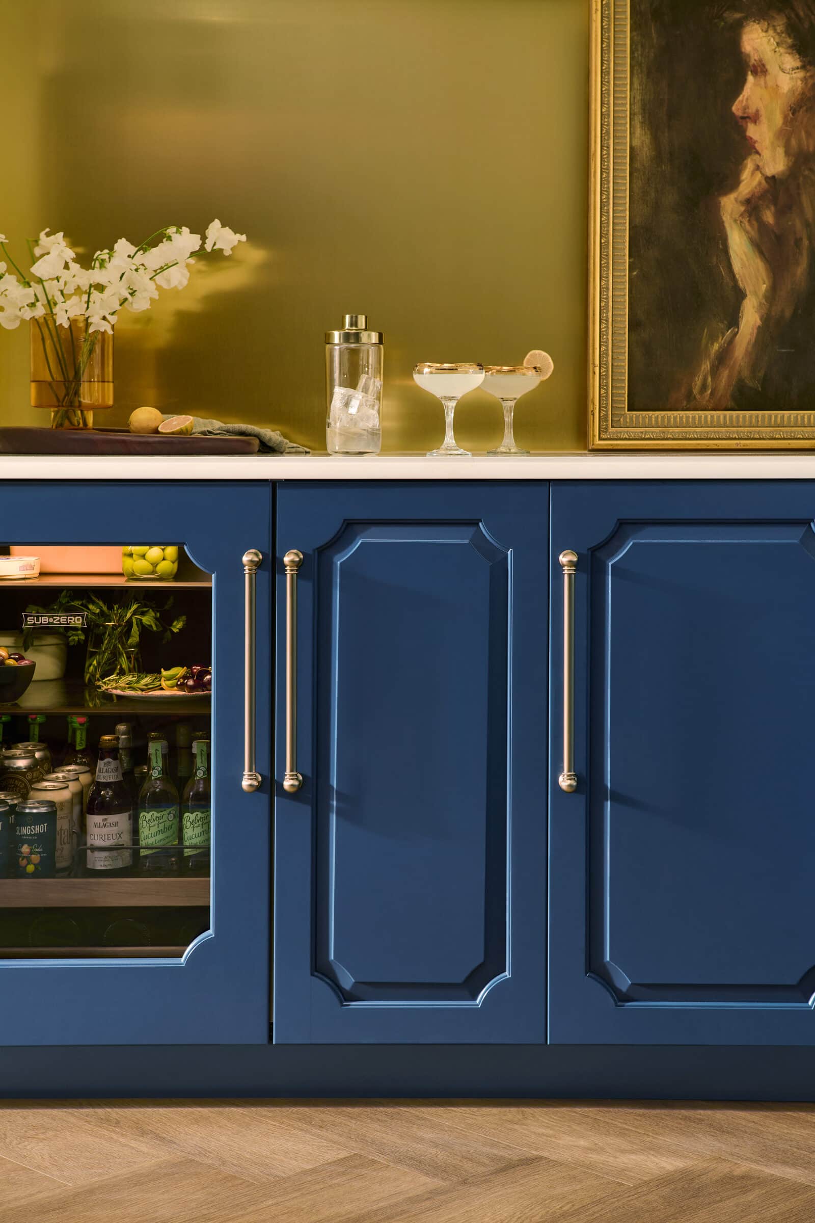 A Sub-Zero glass-door undercounter refrigerator stocked with drinks and produce beside a closed cabinet, set into deep blue bar cabinetry with a brass wall behind