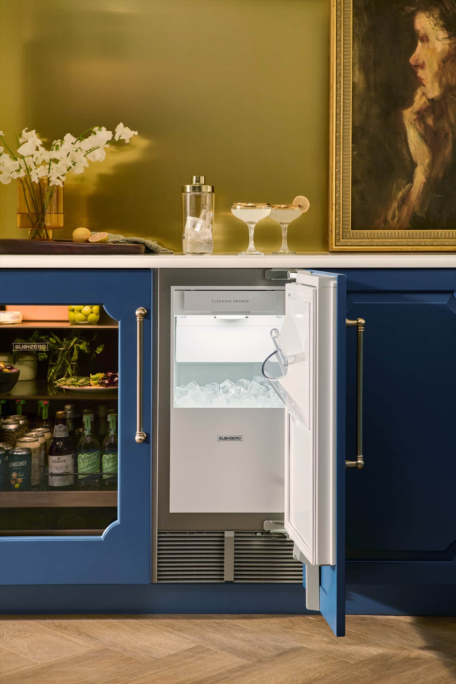 An open Sub-Zero undercounter ice maker filled with ice beside a glass-door Sub-Zero refrigerator in a navy bar with a brass backsplash and a framed portrait above