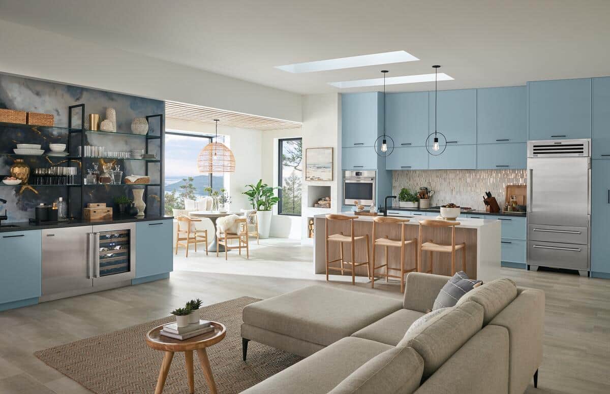 Wide open-plan living room and kitchen with blue cabinetry; a stainless Sub-Zero refrigerator; an undercounter wine unit; and a linen sectional sofa in the foreground