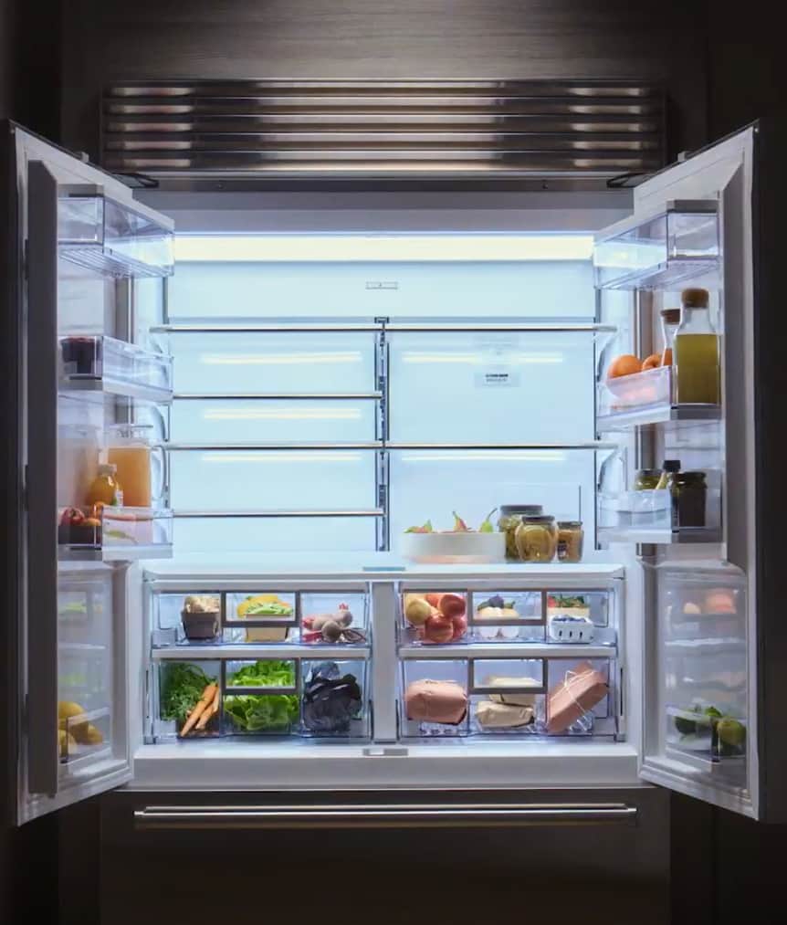 Built-in Sub-Zero refrigerator with both French doors open, showing lit glass shelves and drawers stocked with fresh produce, dairy, and preserves