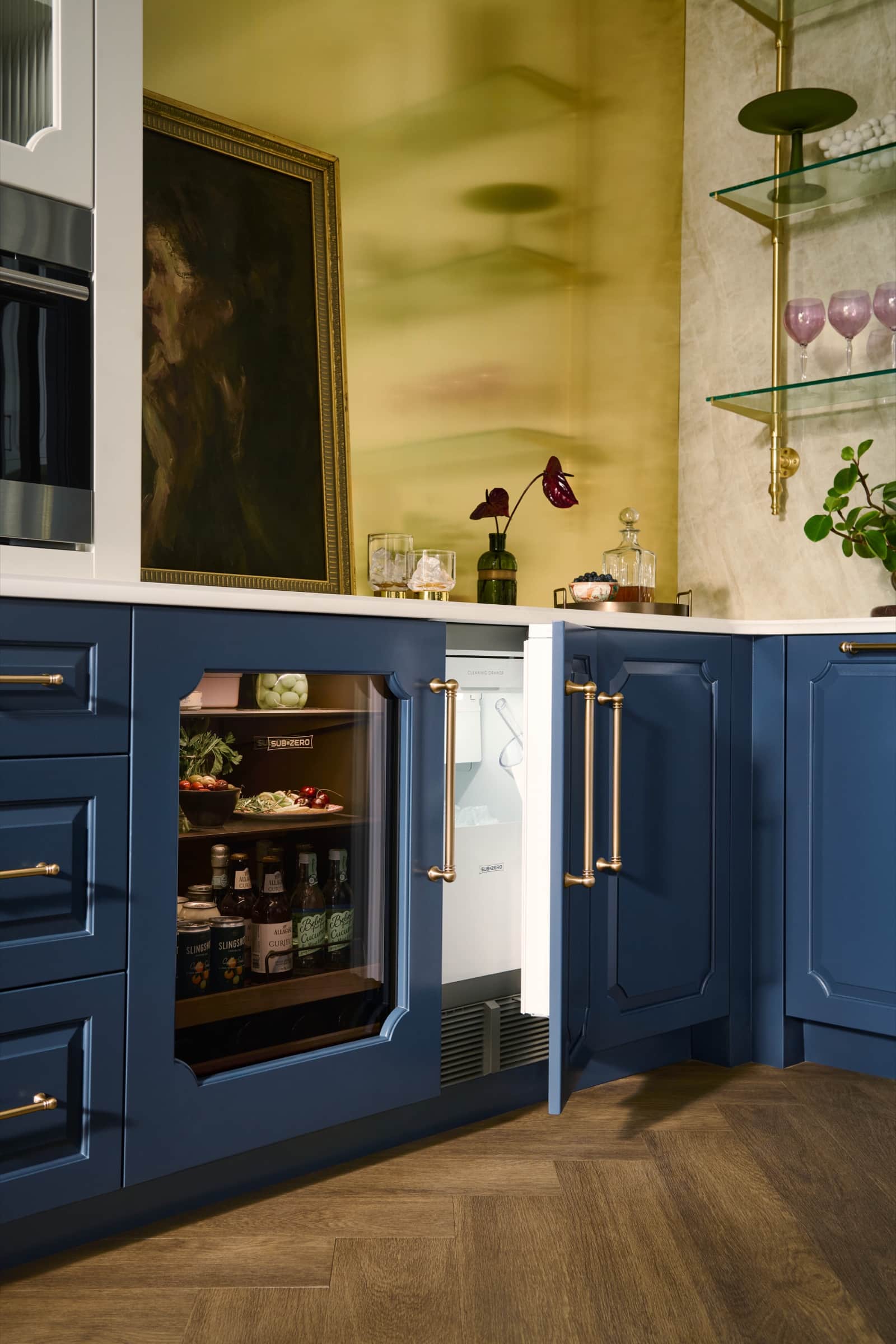 A Sub-Zero undercounter refrigerator and glass-door beverage unit with brass handles set into navy cabinetry in a bar with a brass backsplash