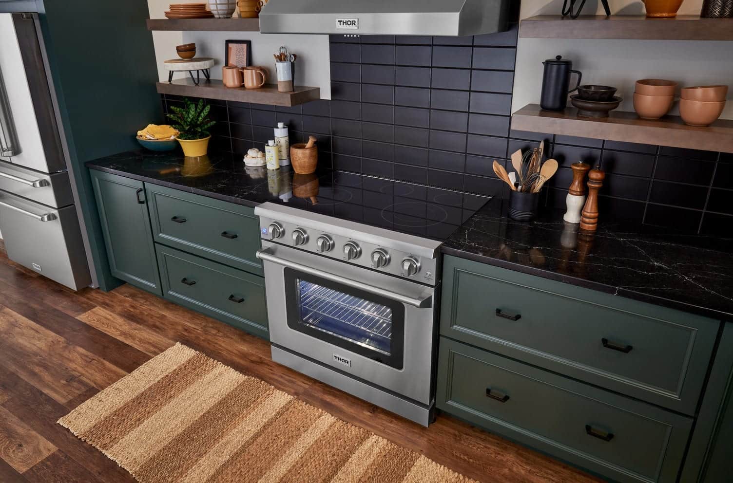 A stainless Thor Kitchen electric range with a black glass cooktop under a stainless Thor Kitchen wall hood, set between dark green cabinets against a black tile backsplash