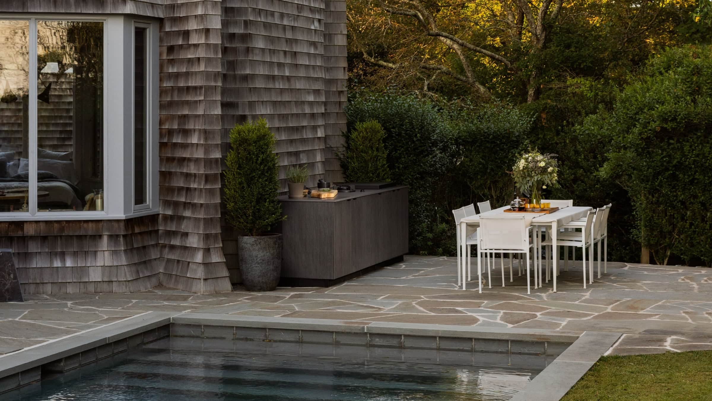 A dark fluted Urban Bonfire outdoor kitchen cabinet with a griddle against a weathered shingle wall, on a stone terrace beside a pool and a white dining table set for six