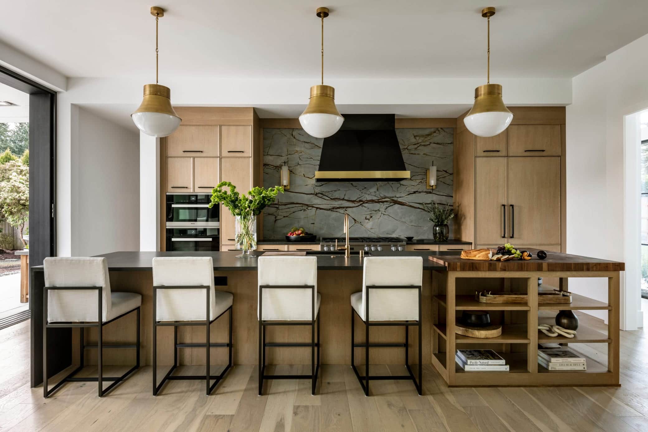 A black Vent-A-Hood wall hood with a brass band against a veined stone slab, above a long oak island with four upholstered stools under brass pendants