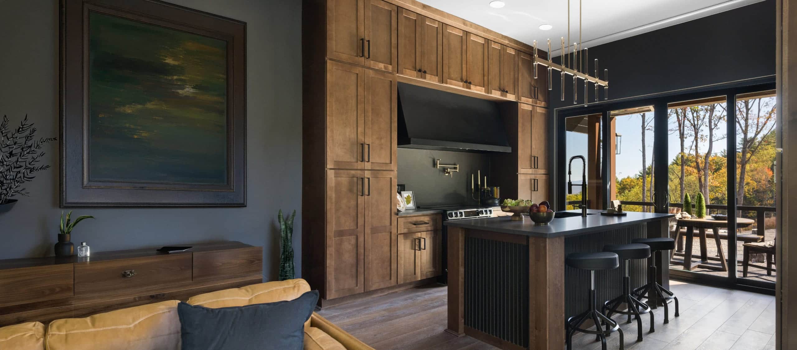 A black Vent-A-Hood wall hood set into an alcove of stained wood cabinetry, a dark island with three stools and glass doors onto a wooded deck