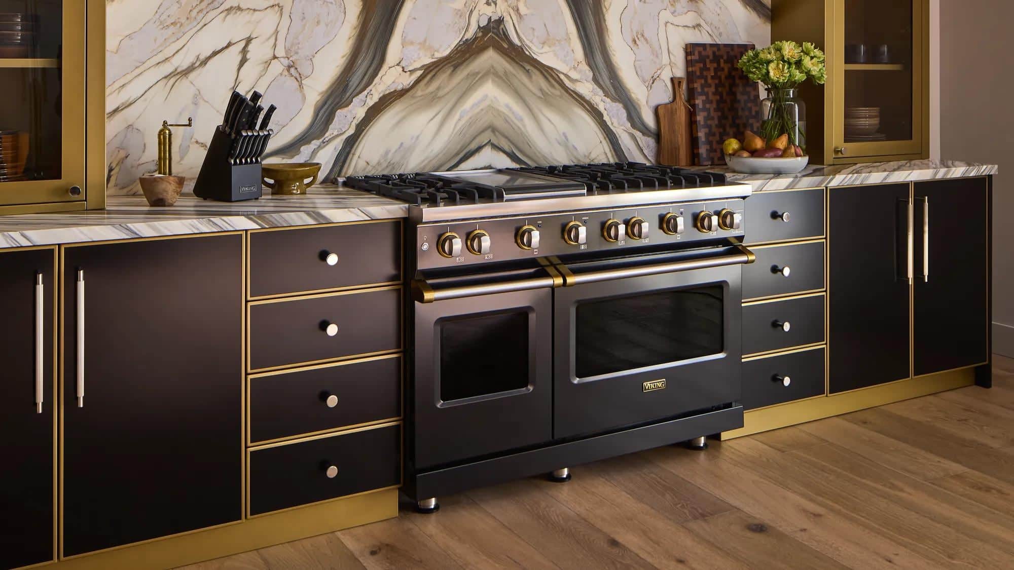 A black Viking range with brass trim and a griddle, set between black and brass cabinetry against a figured marble slab backsplash