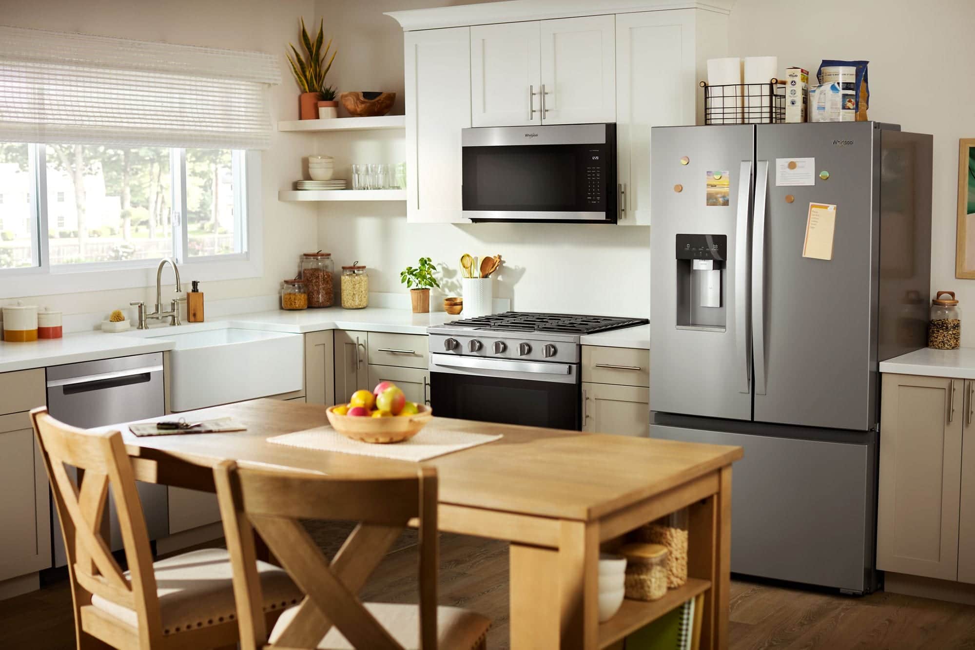 A stainless Whirlpool gas range, over-the-range microwave and French-door refrigerator in a light kitchen with a farmhouse sink and a wood table in front