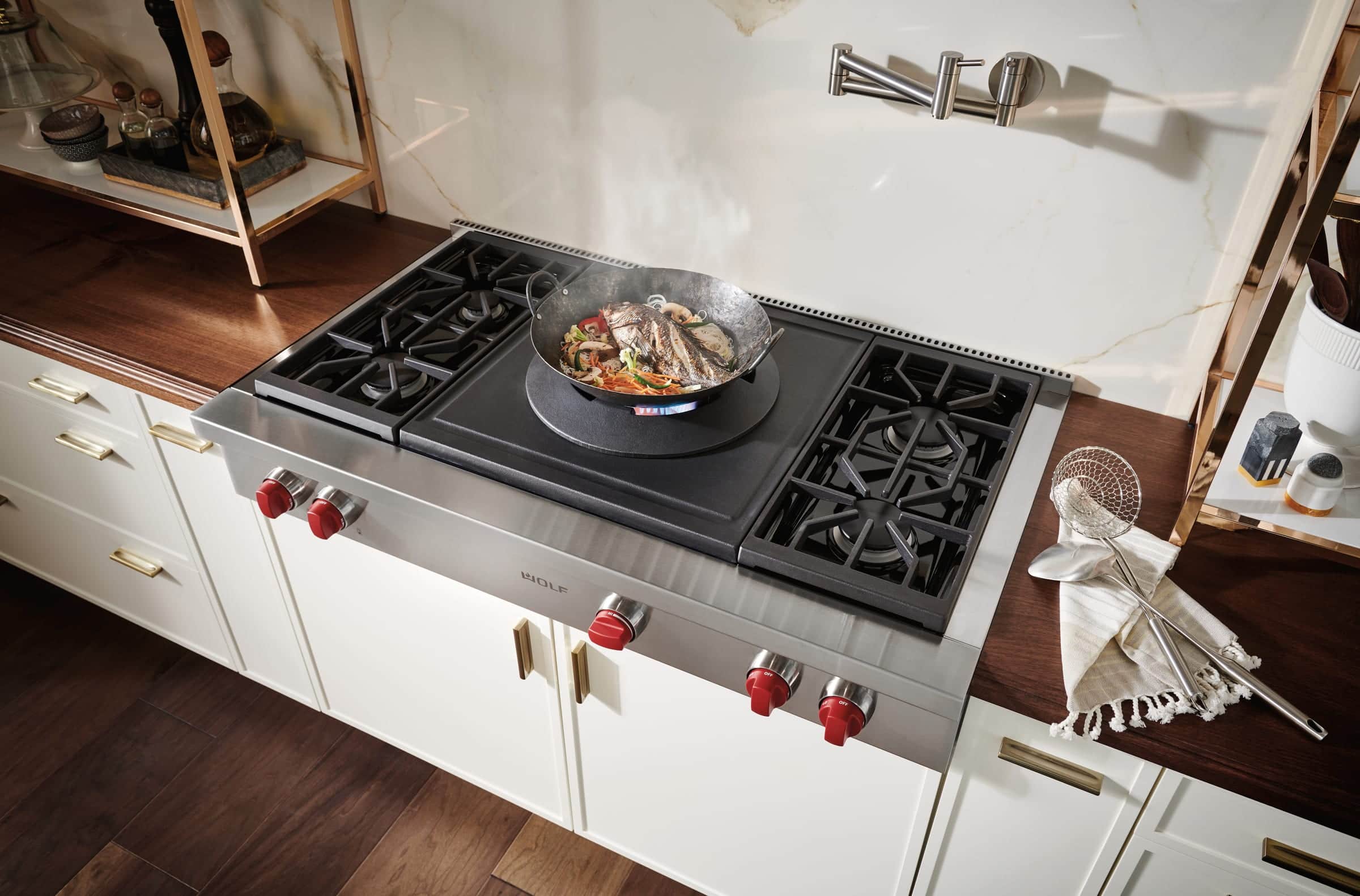 A Wolf stainless gas rangetop with red knobs, a wok of fish and vegetables over a blue flame, a griddle and two more burners, set in white cabinetry under a marble backsplash