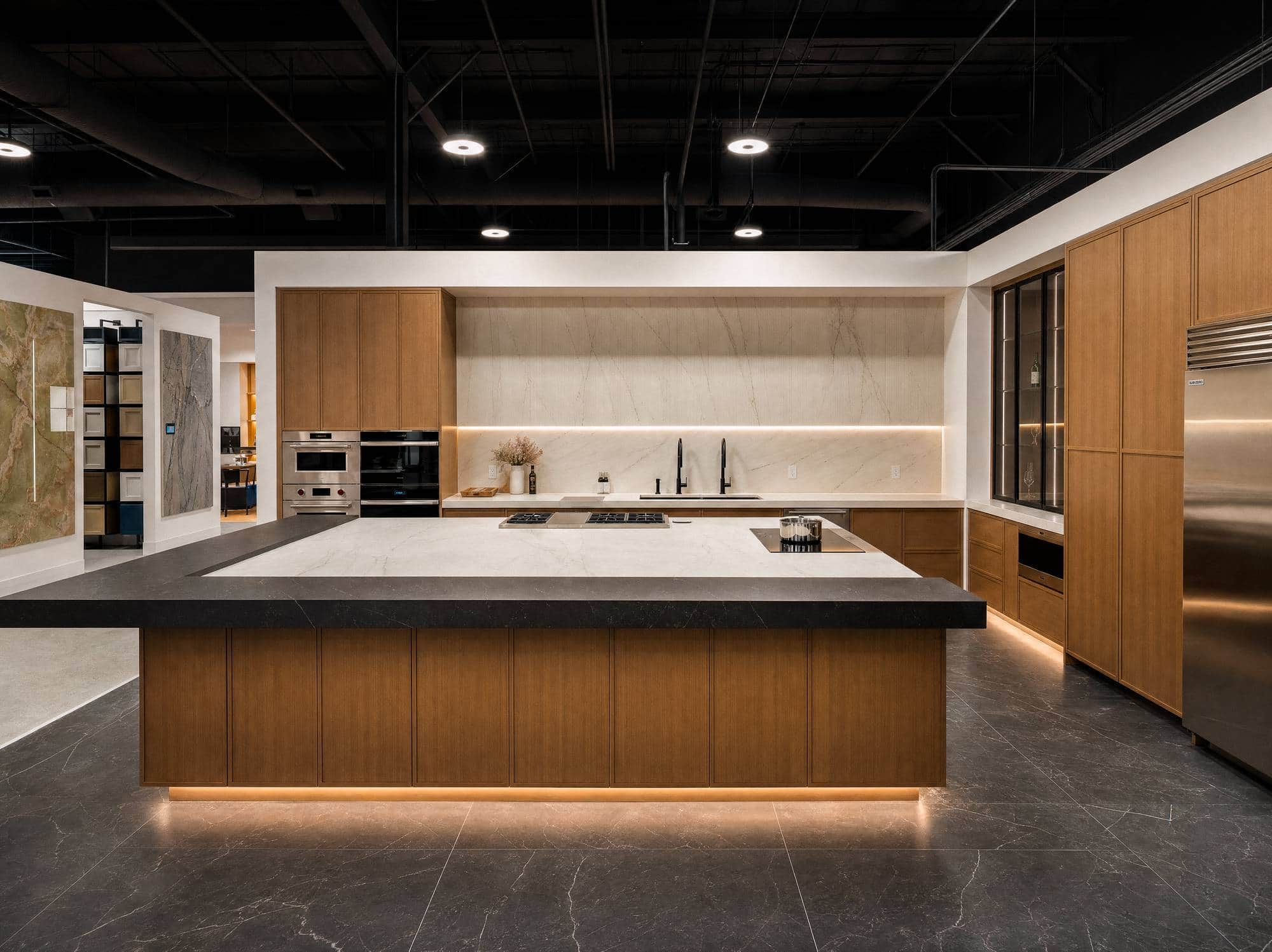 The Roseville showroom floor: a walnut island with a cooktop under a dark exposed ceiling, wall ovens and refrigerator columns receding into the distance