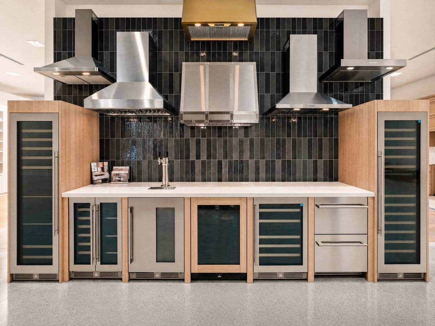 A display wall of five ventilation hoods over rangetops, black tile behind, wine columns flanking