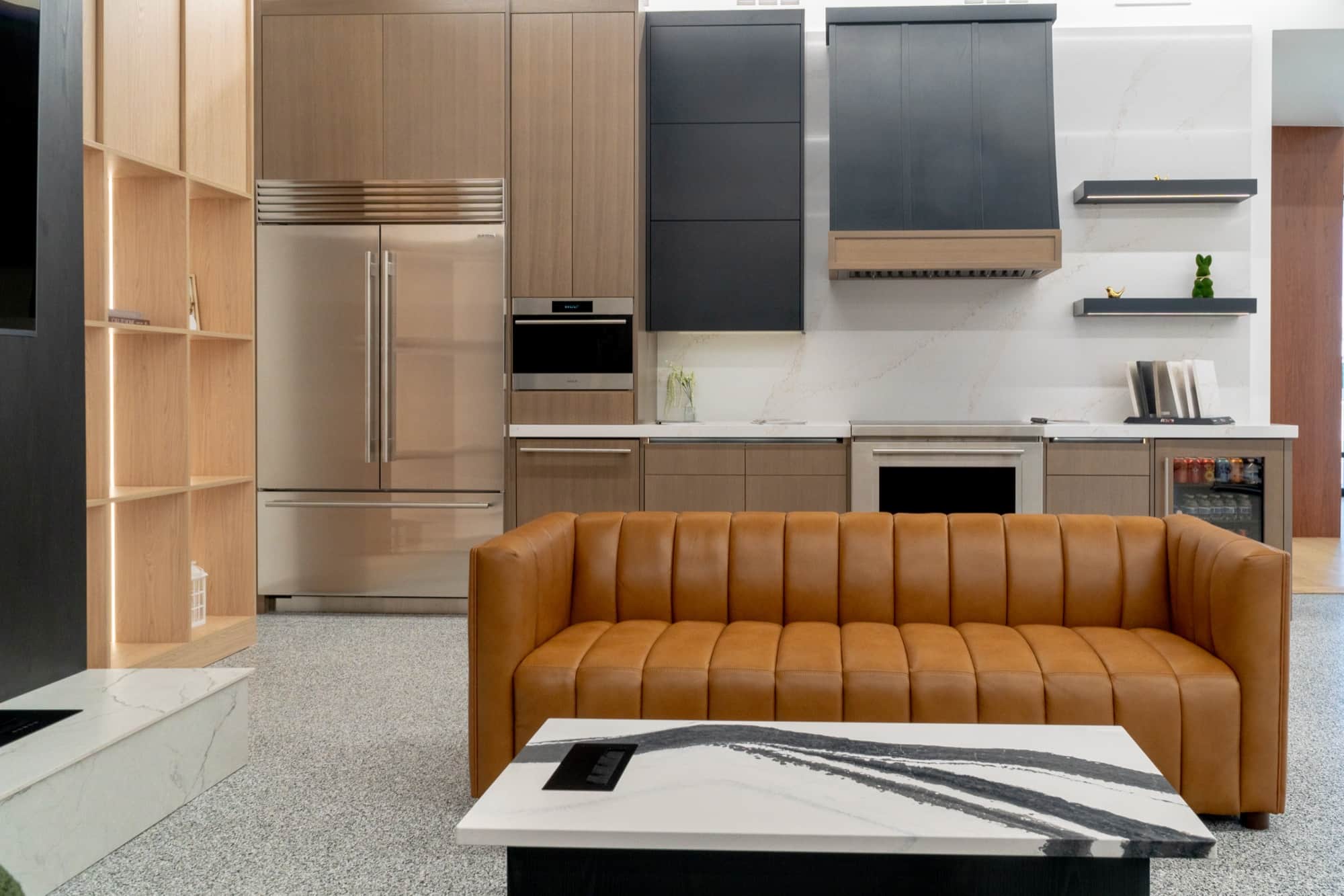 A kitchen vignette on the Post Falls showroom floor with a panel-ready Sub-Zero column refrigerator and a Wolf wall oven set into oak cabinetry, a wood and black chimney hood over the range, and a leather sofa in the foreground