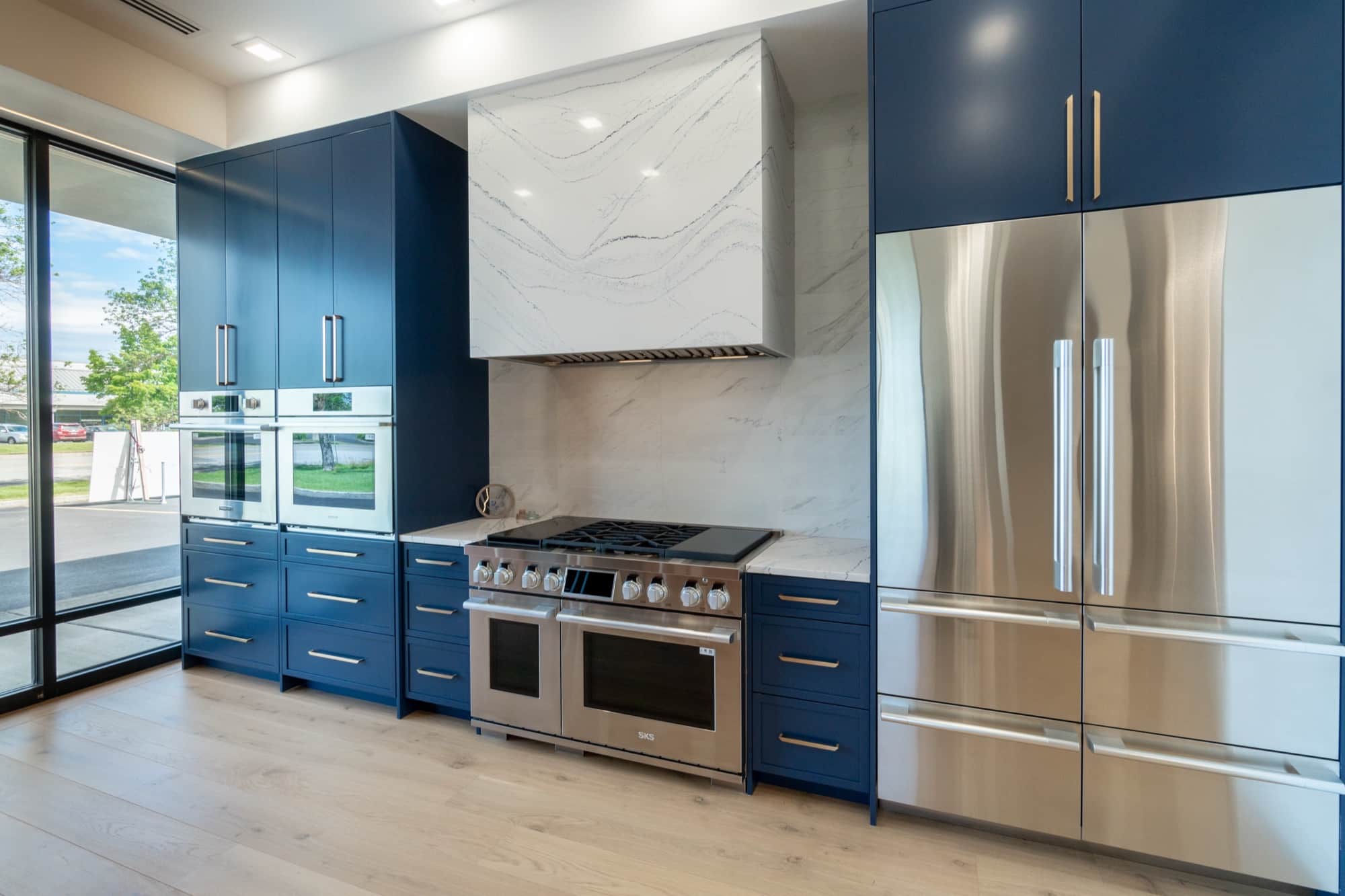 A full kitchen vignette bay with navy cabinetry, two stainless wall ovens, and an SKS professional dual-oven range with a griddle under a quartz-clad chimney hood
