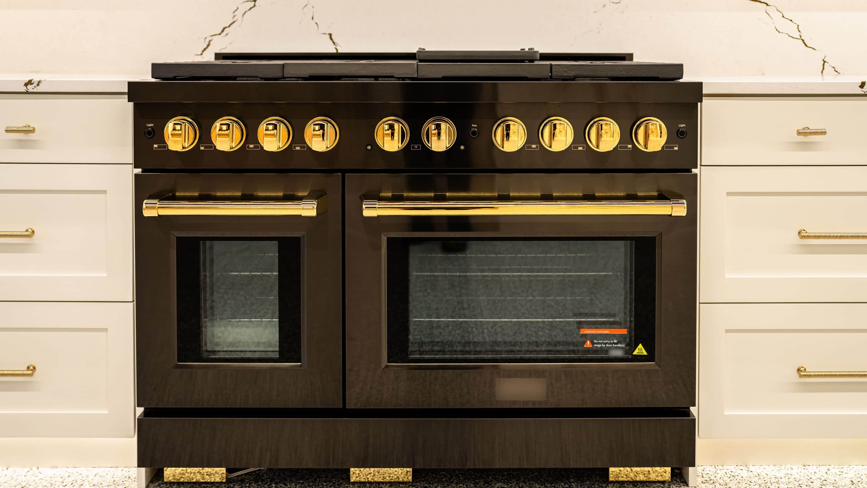 A black dual-oven range with brass knobs and brass handles between white shaker drawer runs under a gold-veined white slab
