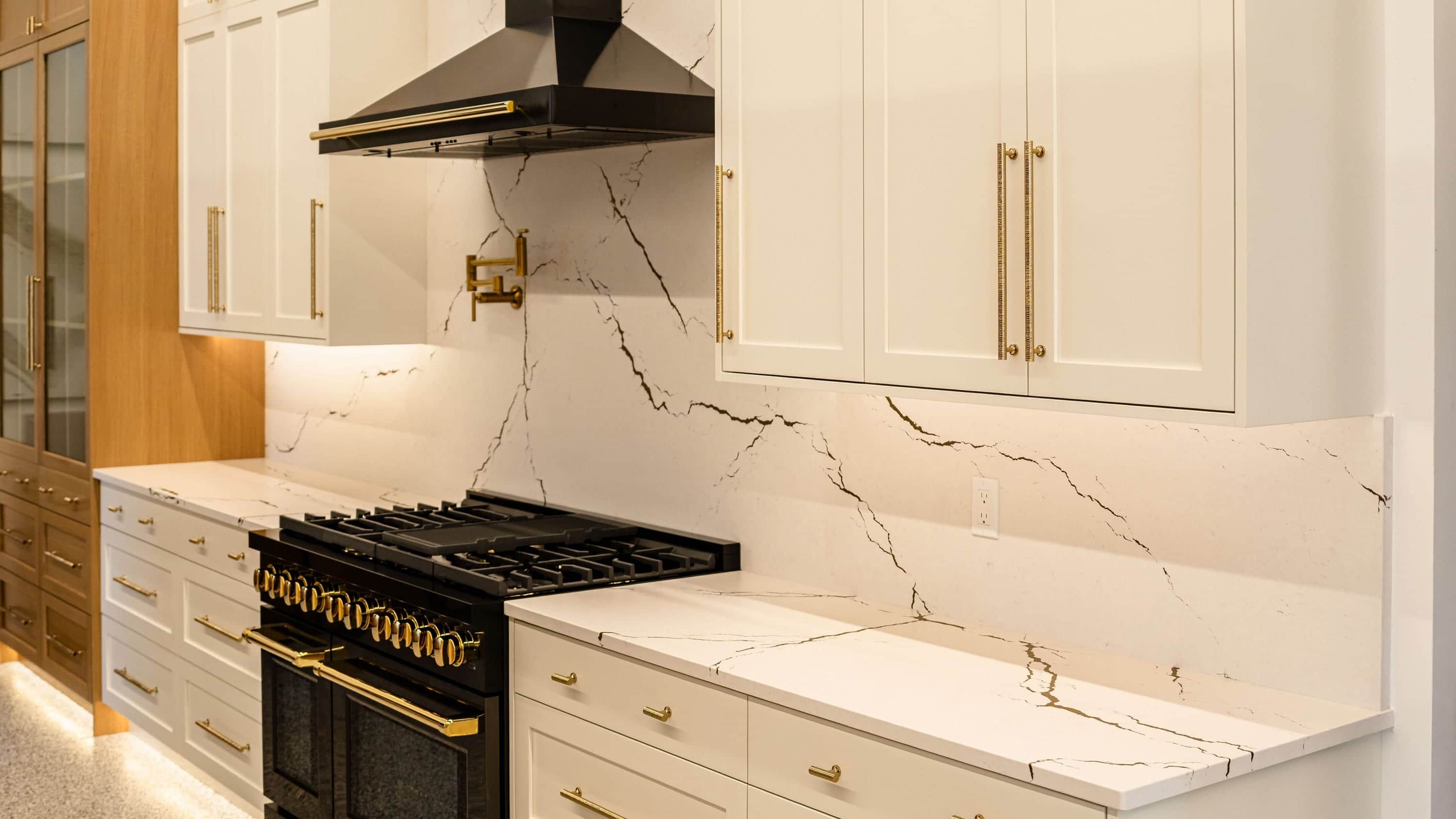 A white shaker kitchen wall with a black chimney hood, a brass pot filler on a gold-veined white slab backsplash and a black range with brass knobs