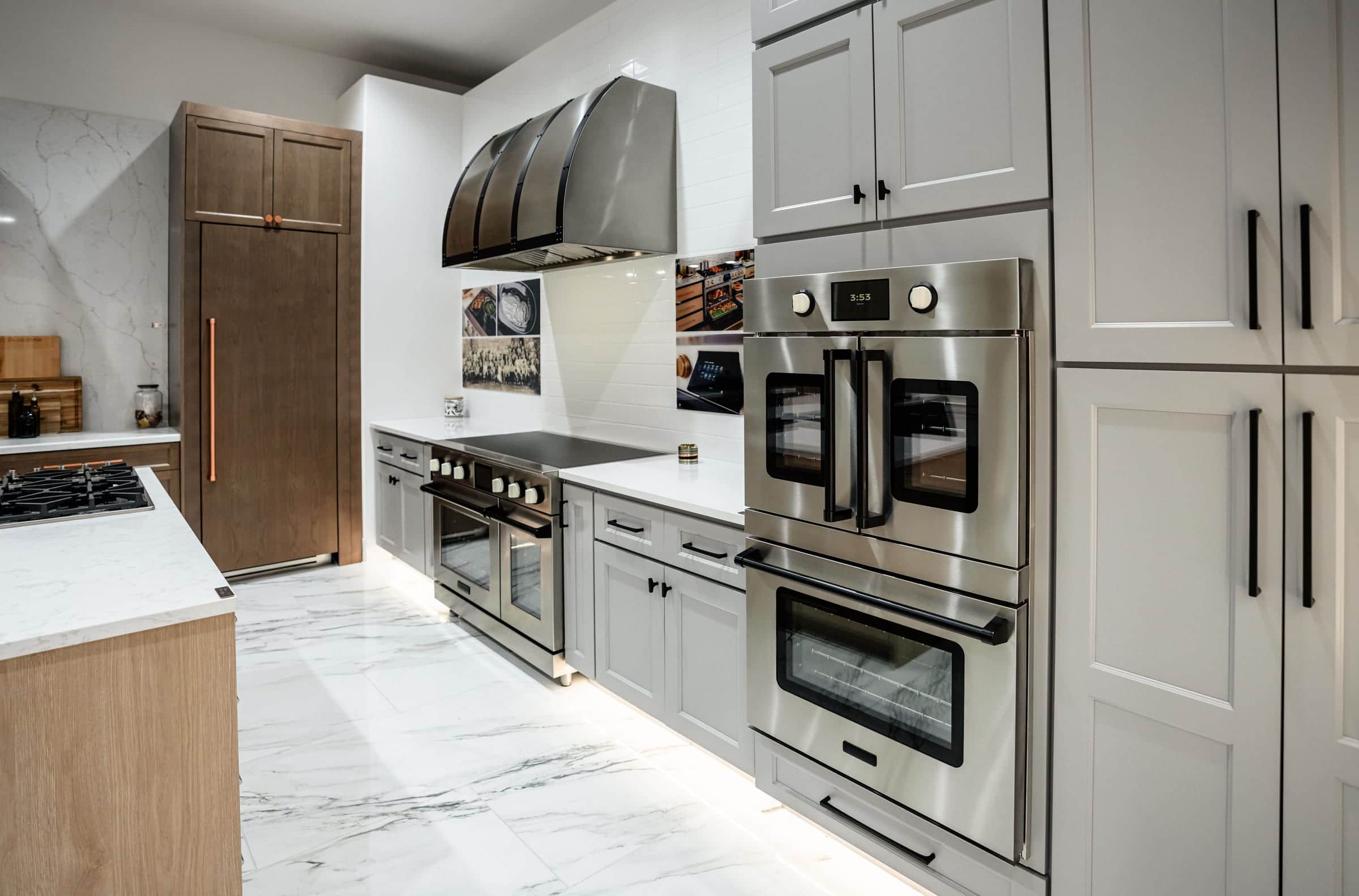 A galley display in the Roseville showroom: an oak island with a gas cooktop, a walnut panel-ready column, a strapped stainless hood over a range and stacked wall ovens set into grey painted cabinetry