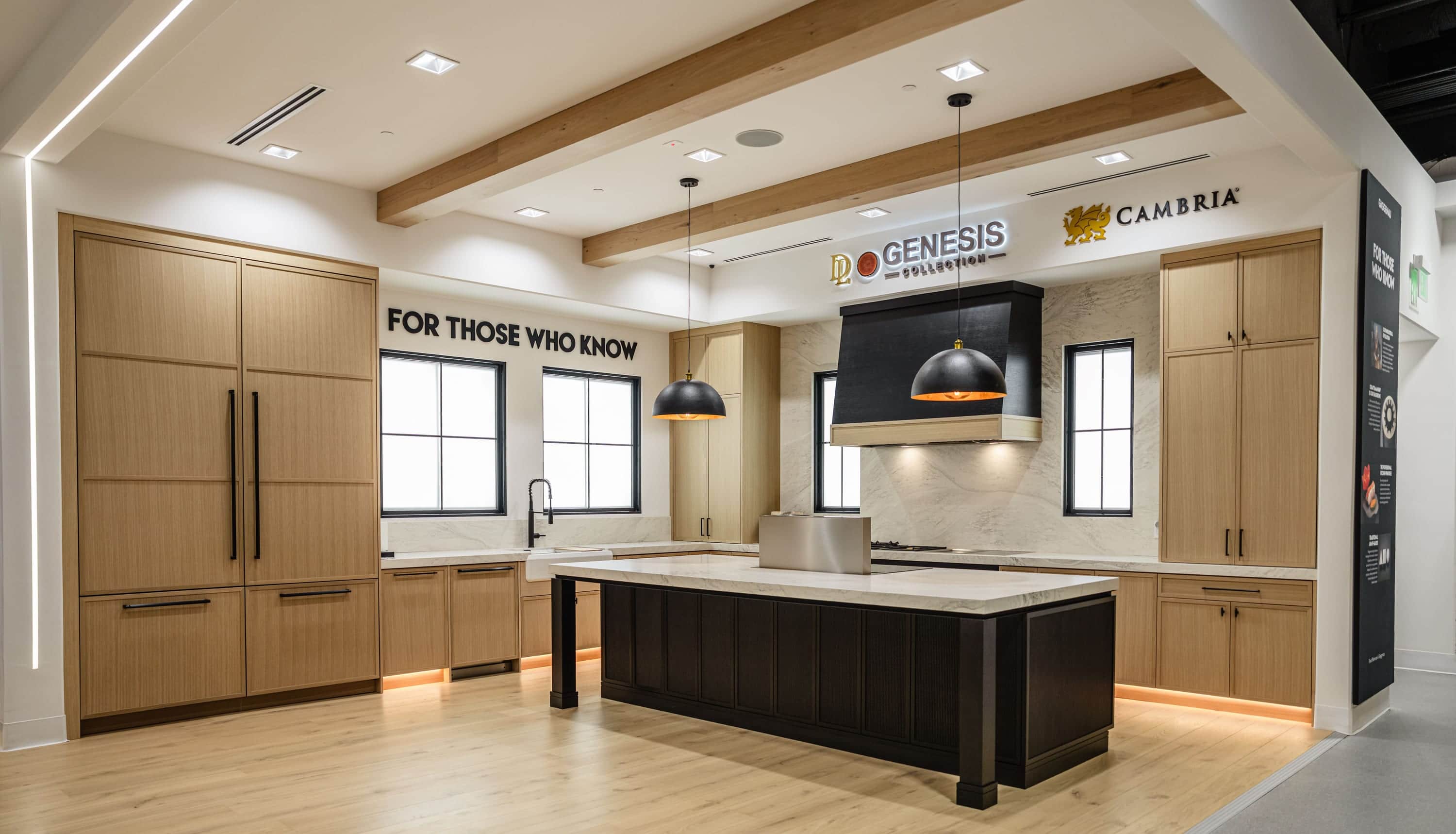 A display kitchen in the Roseville showroom: tall oak cabinetry with a panel-ready column, a black island under two dome pendants and a black hood over a stone backsplash