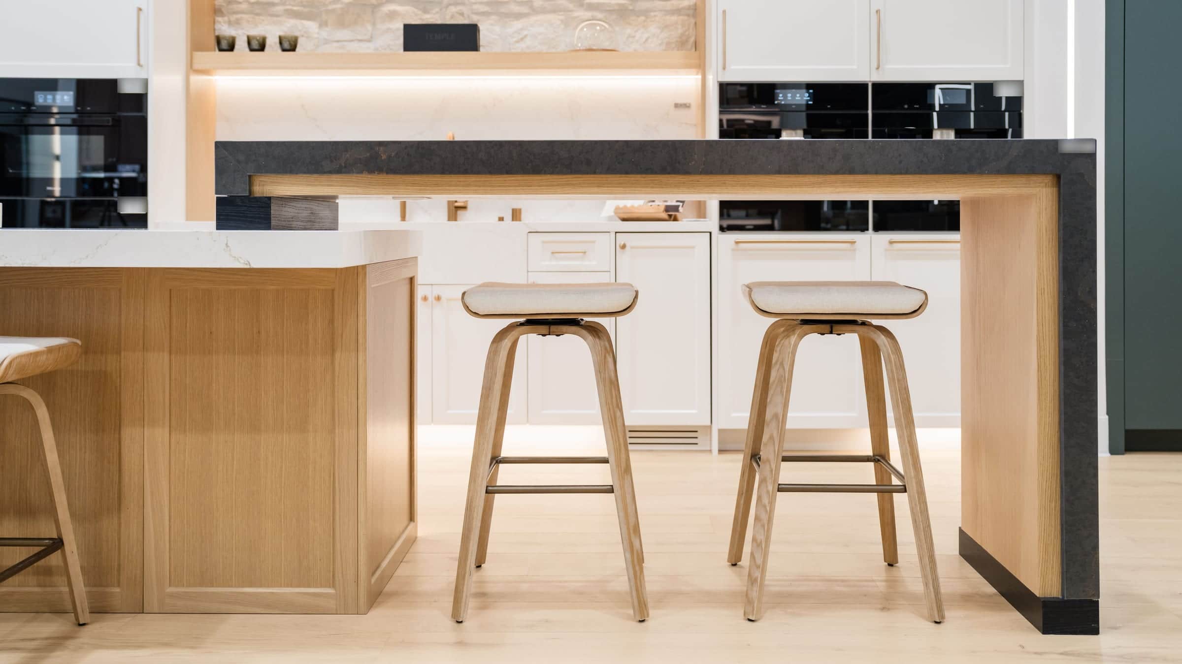 Two wood bar stools under a dark stone peninsula on a rift oak island in the Roseville showroom, with white cabinetry and built-in coffee systems behind